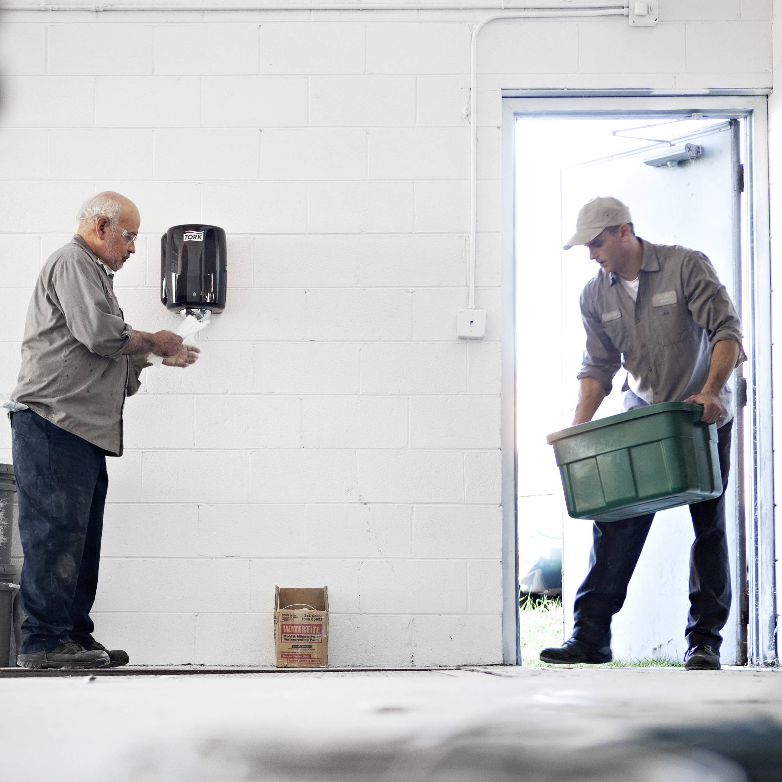 Two men in workwear: One is carrying a box through an open door, the other is using a paper towel dispenser on the wall.