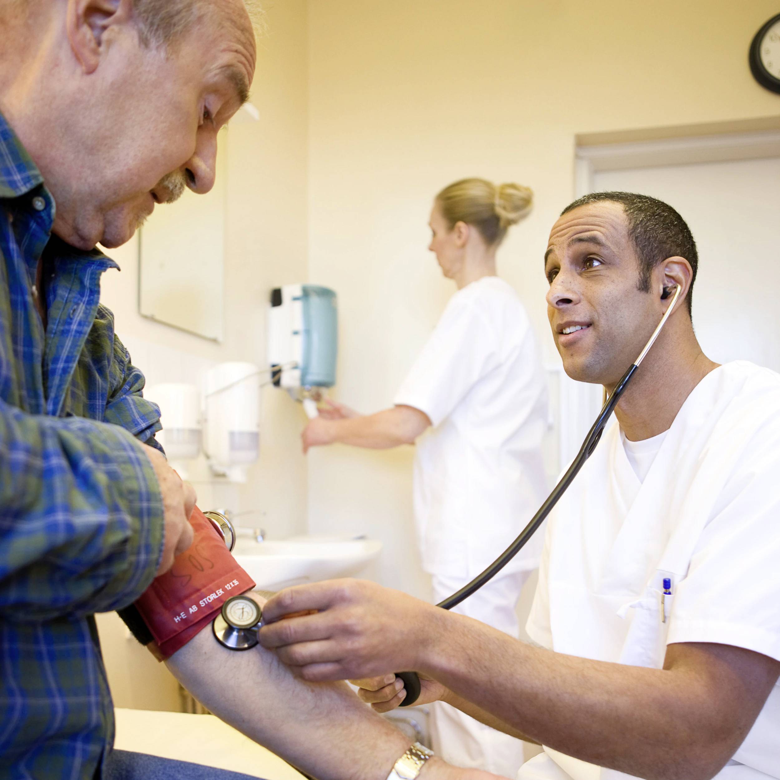 A doctor measures the blood pressure of an elderly man in a clinic. Another person in the background is operating medical equipment.