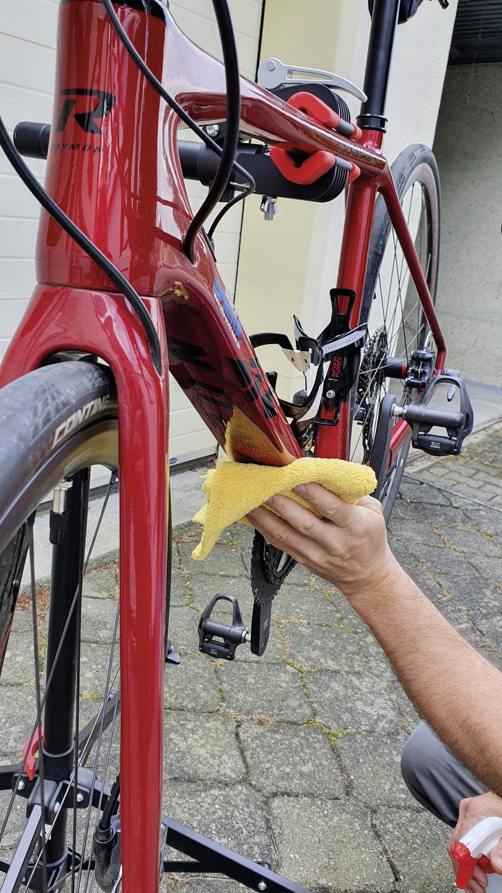 A person is polishing a red bicycle with a yellow cloth while it stands on a bicycle stand. A garage door is visible on the left.