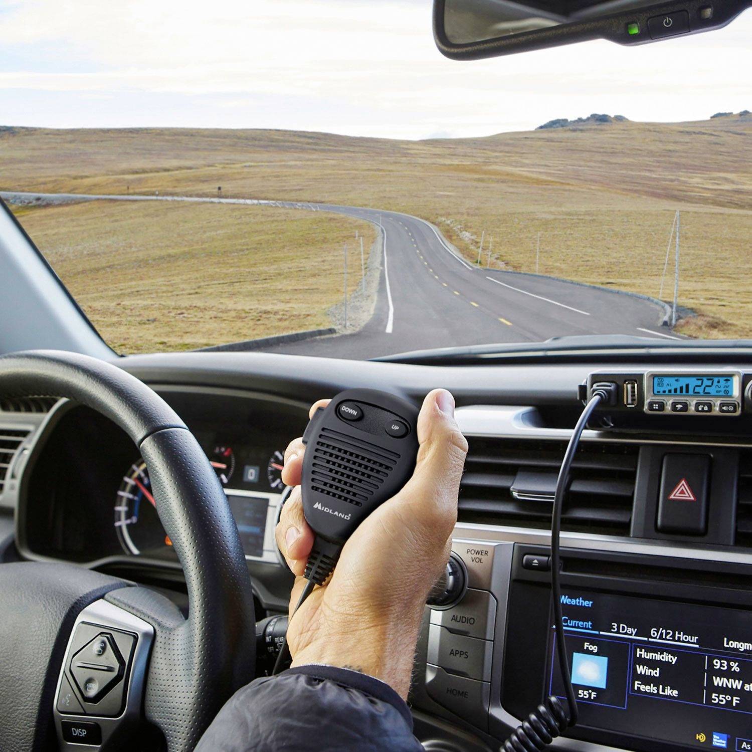 View from a car window of a winding road. One person is holding a walkie-talkie. Traffic-free, rural landscape in the background.