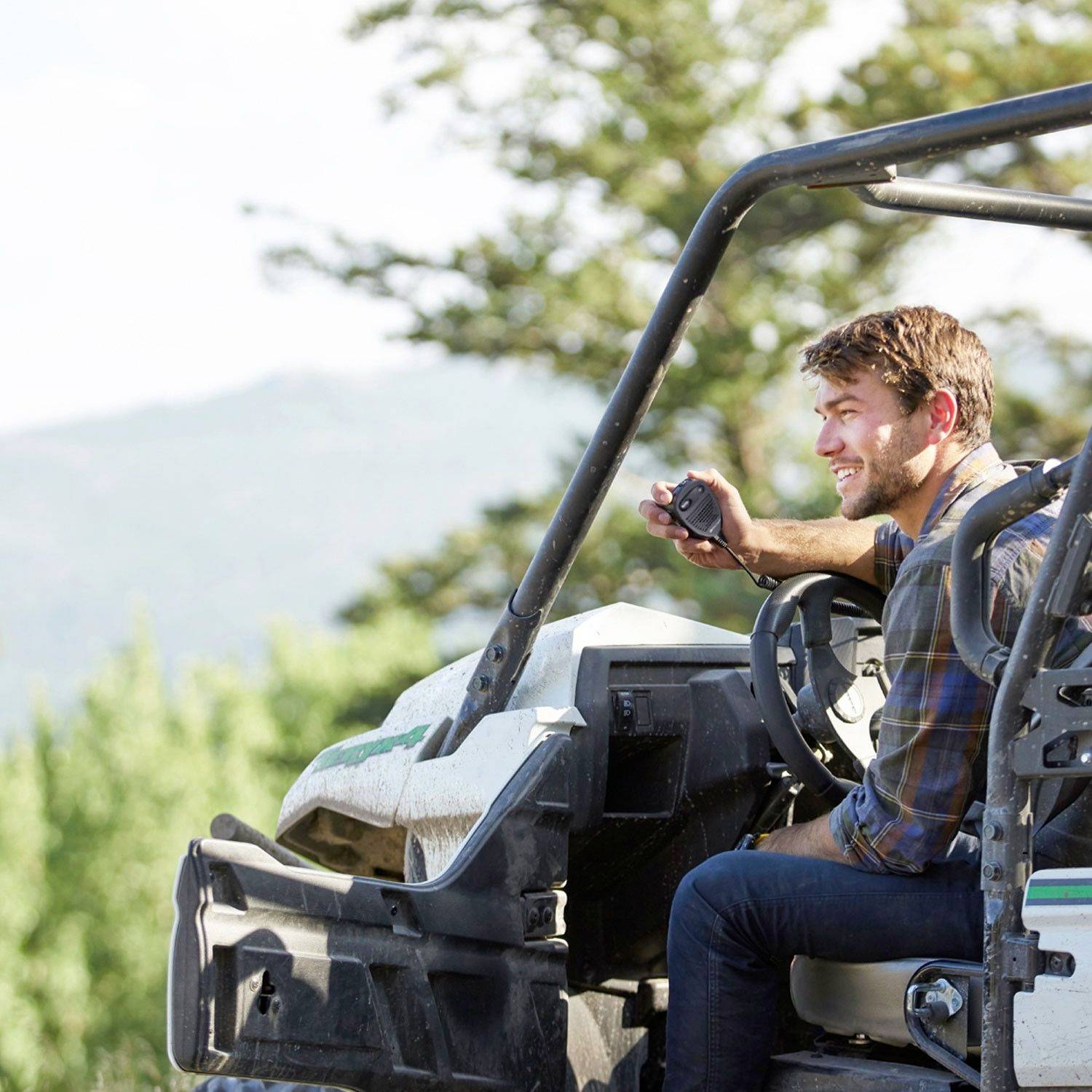 A man is sitting smiling on a quad bike in a natural setting, surrounded by trees and hills. He is holding a camera in his hand.