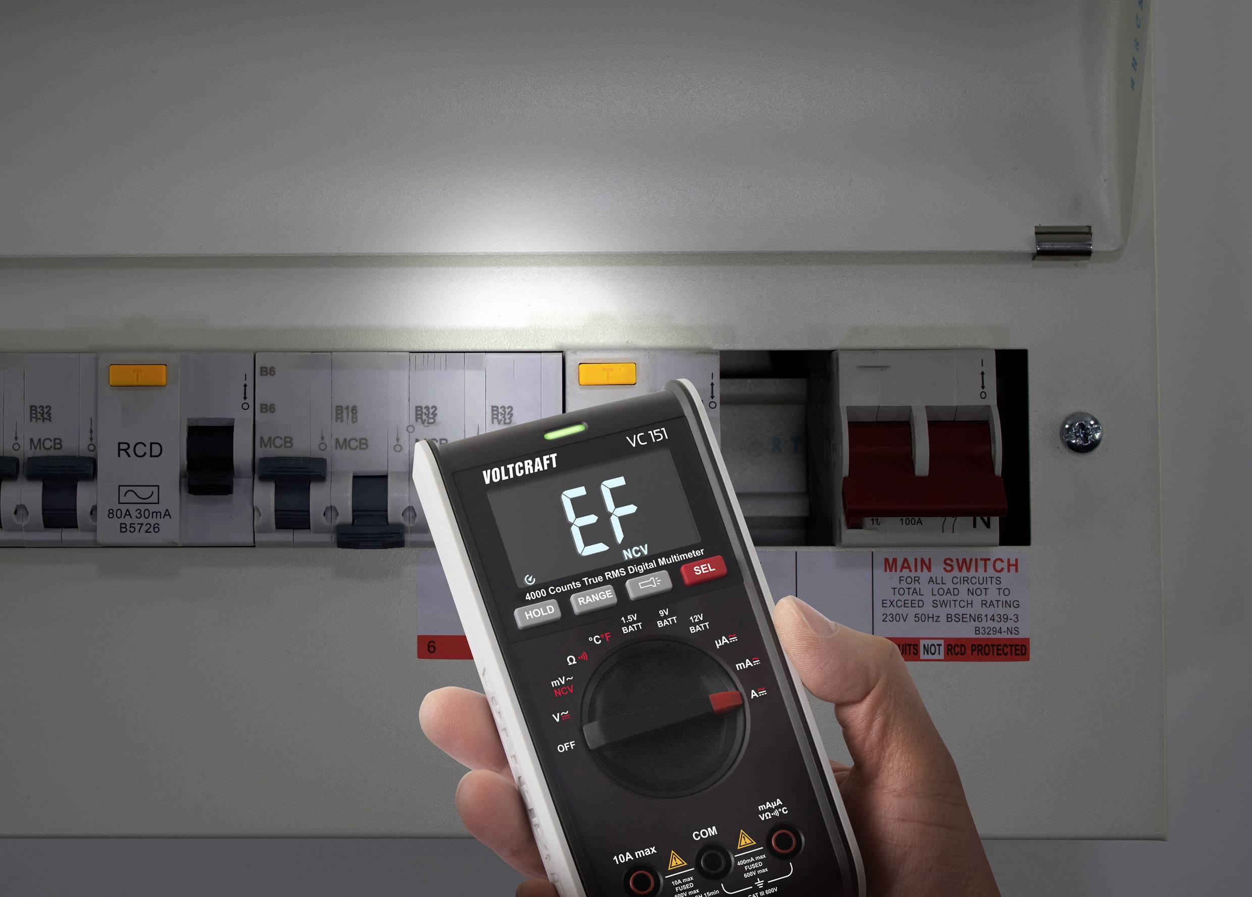 A hand holds a digital multimeter displaying 'EF' in front of an open electrical cabinet with protective switches and main switch.