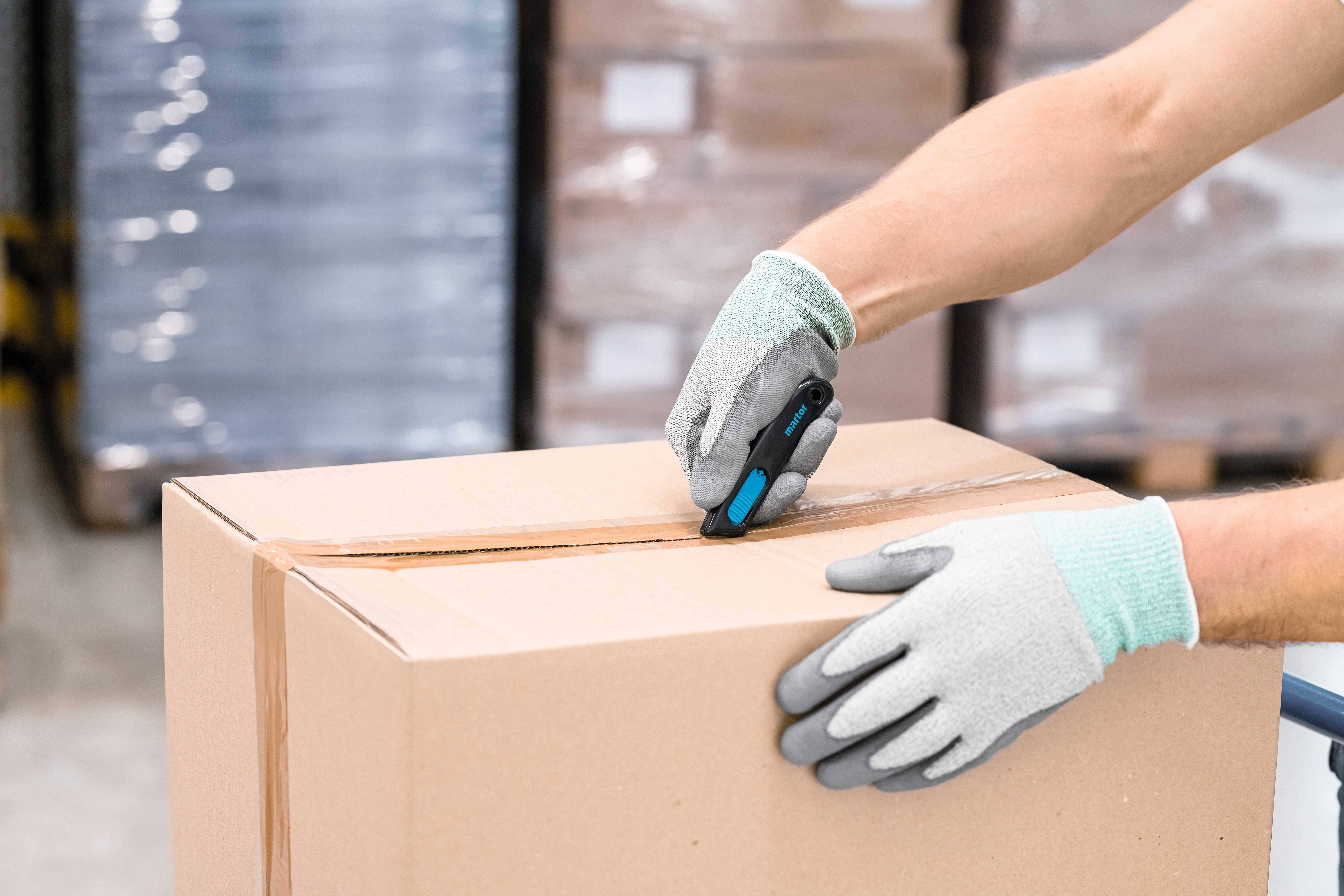 A person wearing gloves is cutting cardboard with a knife. The background shows stacked pallets wrapped in plastic.