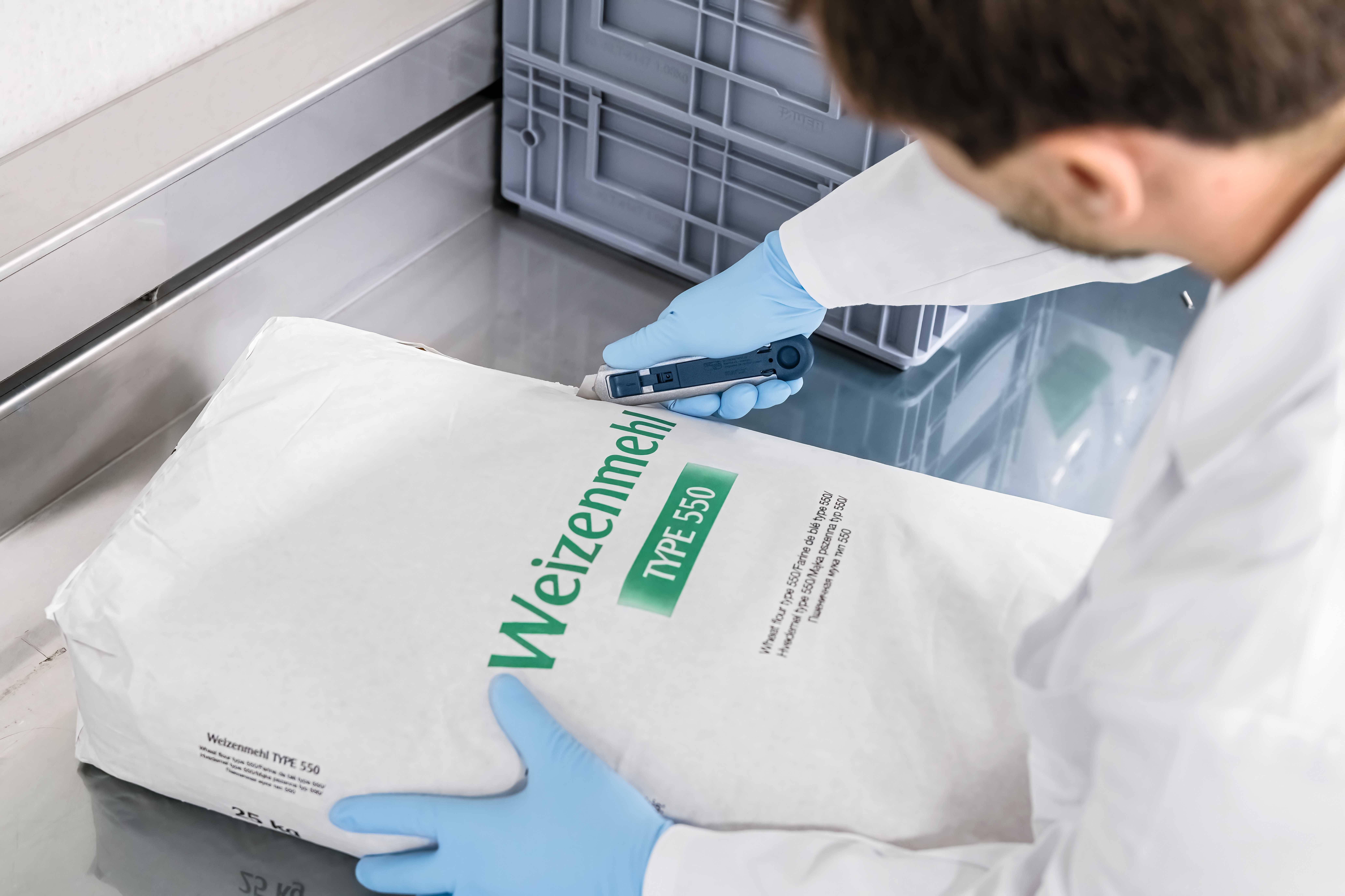 A person in a white lab coat is examining a flour package labelled 'Wheat Flour Type 550' on a stainless steel table.