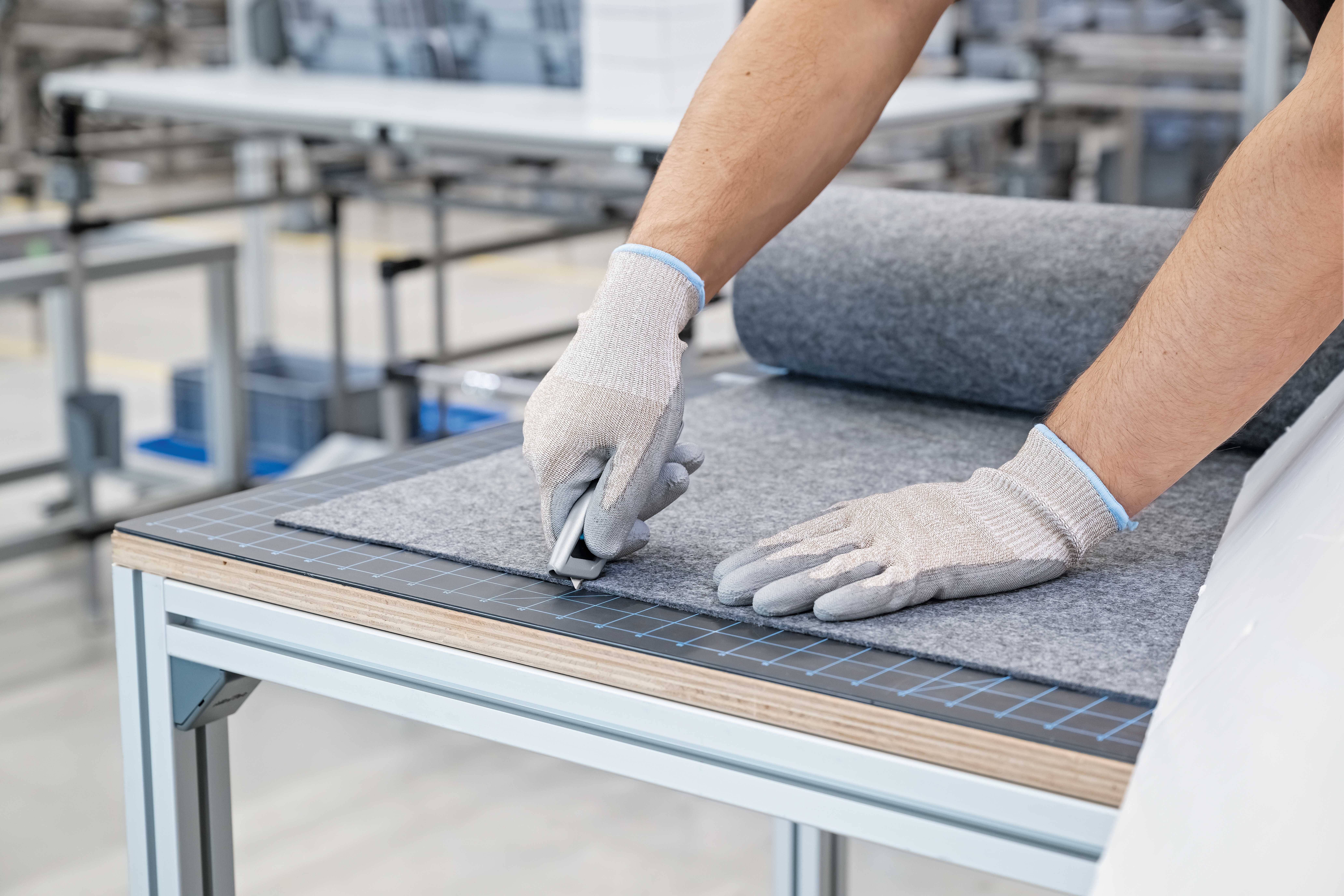A person wearing gloves is cutting grey felt with a knife on a table in a workshop.