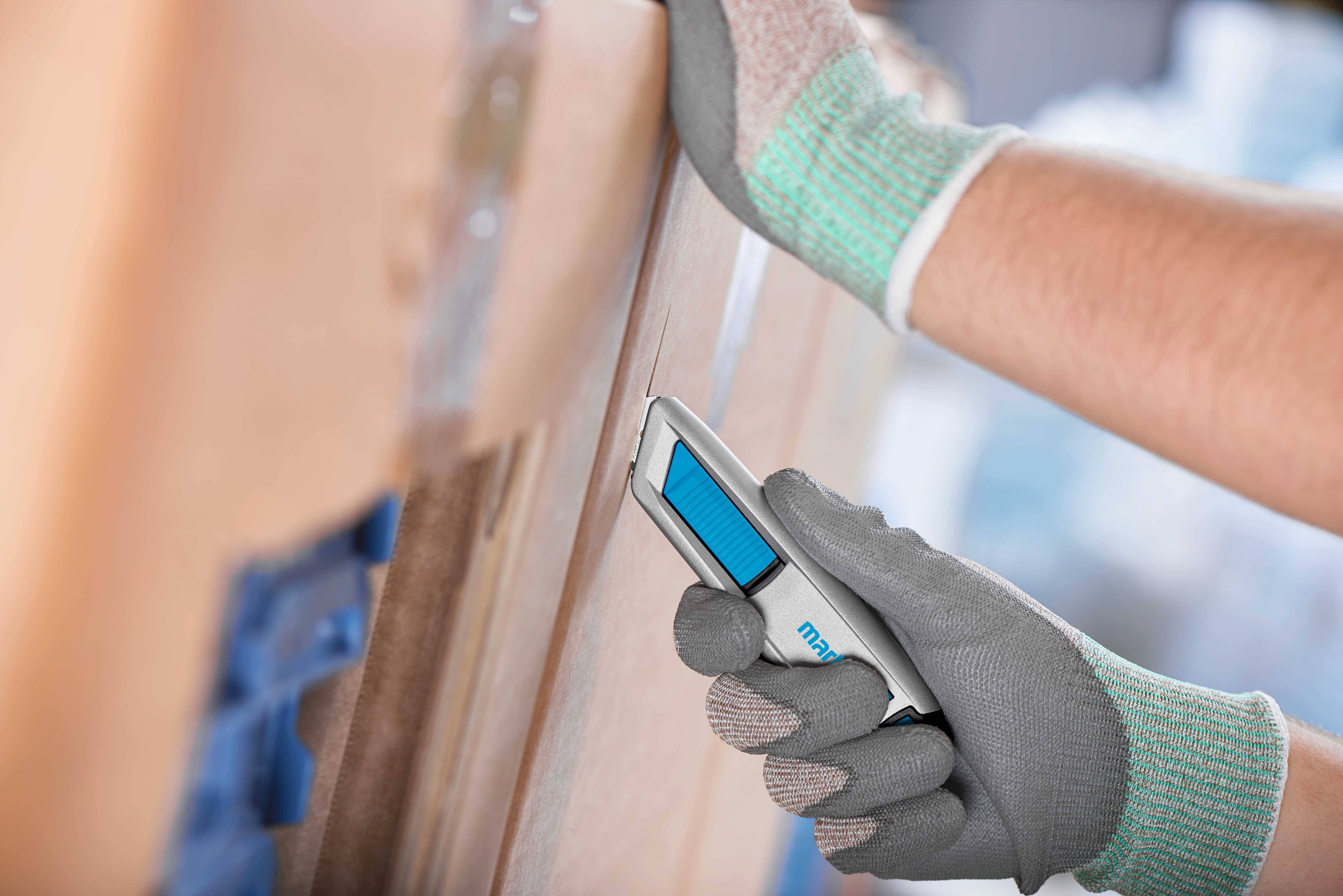 A person wearing work gloves is cutting a cardboard box with a safety knife.
