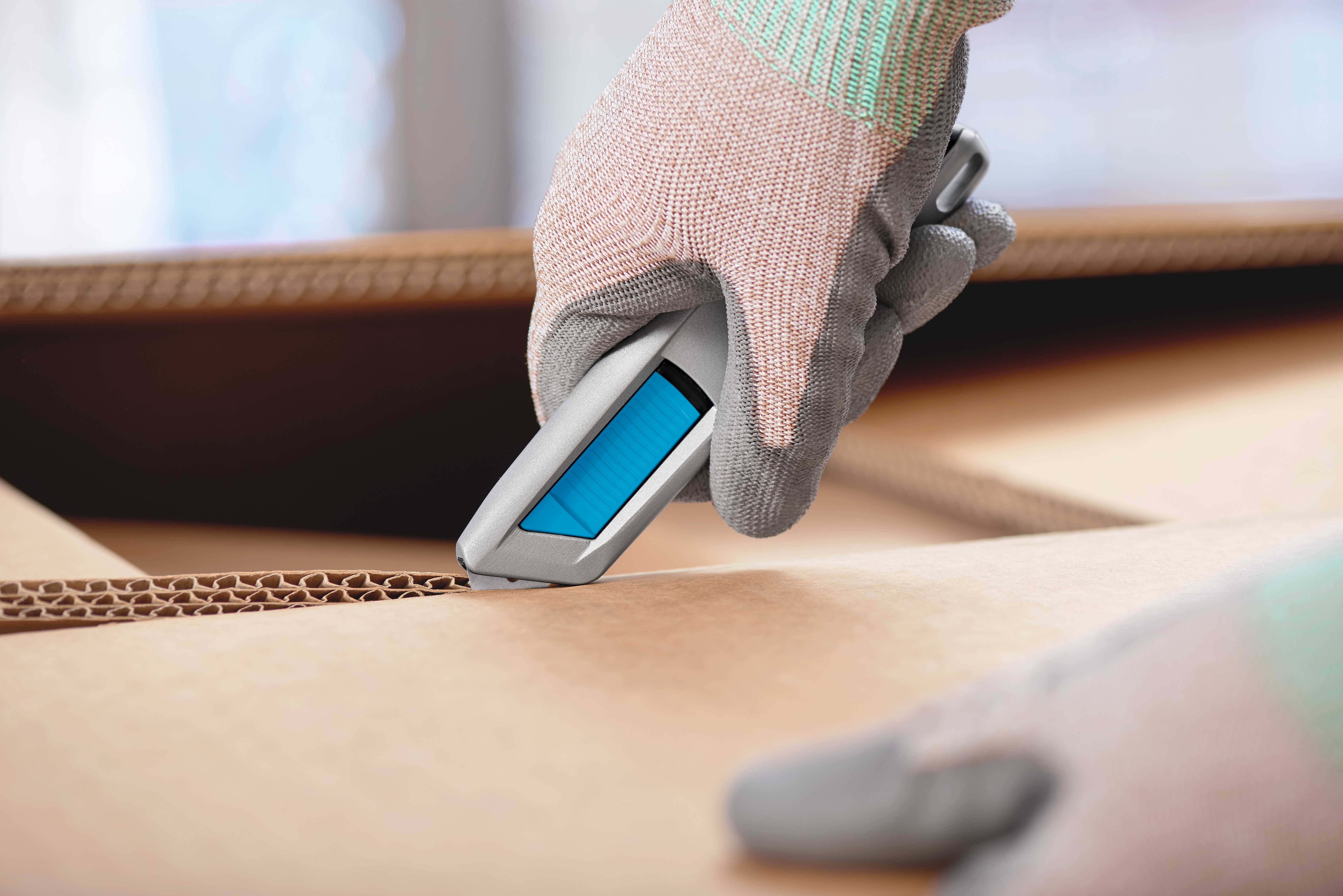 A hand wearing gloves cuts a cardboard box with a utility knife. Further cardboard boxes are blurrily visible in the background.