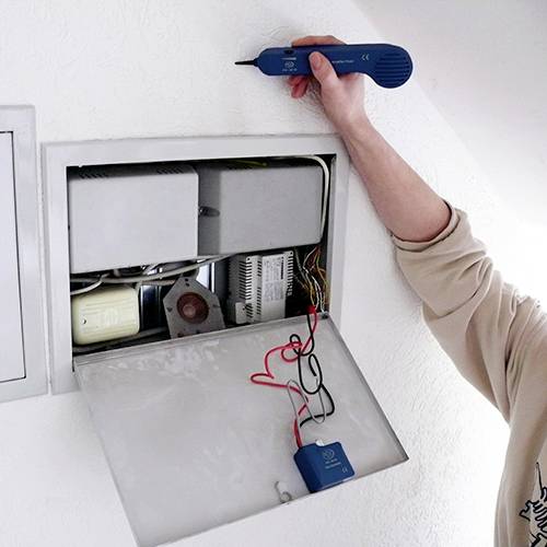 A person is repairing an electronic system in a wall opening using tools. The device is being wired, with various components visible.