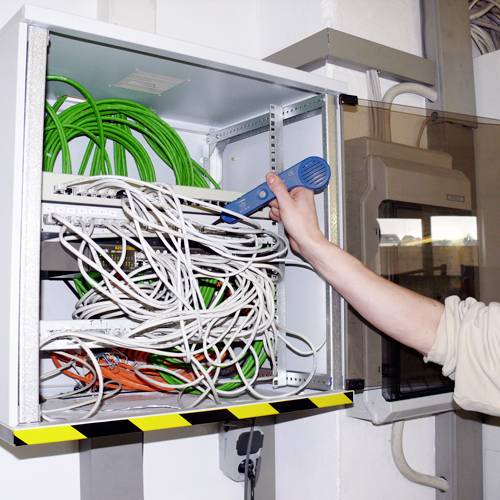 A person is using a blue tool to test a cable in an open control cabinet with many green and white cables.