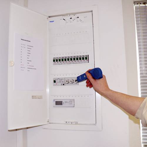 A person is checking the circuits of an open fuse box in an interior space using an electronic device.