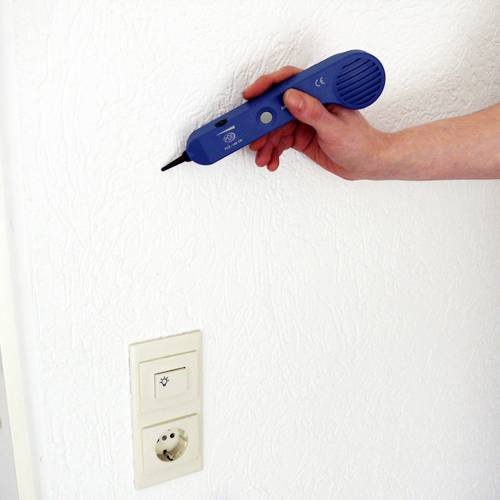 A hand is holding a blue device against a white wall above a power socket.