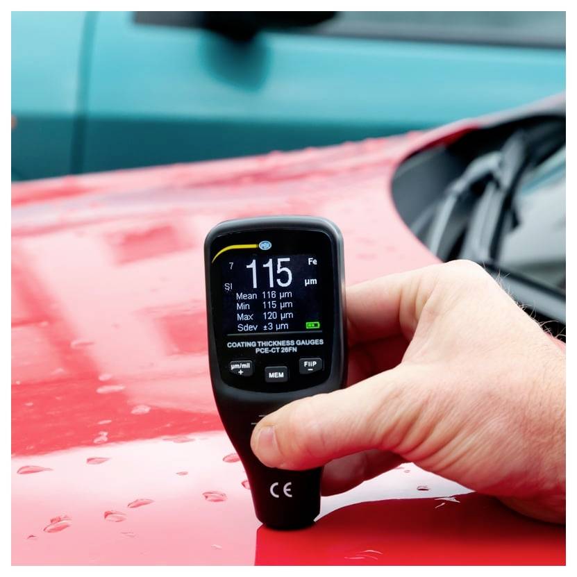 Handheld coating thickness gauge measures paint on a red car hood, showing 115 µm on its display.