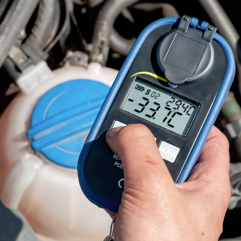 A hand is holding a digital coolant testing device displaying '-33.7°C'. A coolant container is visible in the background.