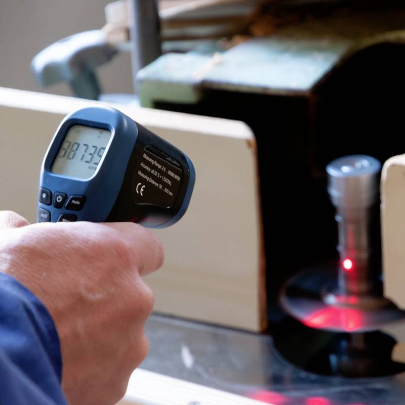 A person is holding an infrared thermometer, reading a temperature of 87.3 °C, apparently measuring a machine.