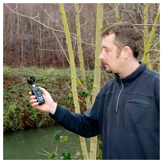 Man outdoors holding a handheld wind meter near a wooded stream, apparently measuring weather conditions.