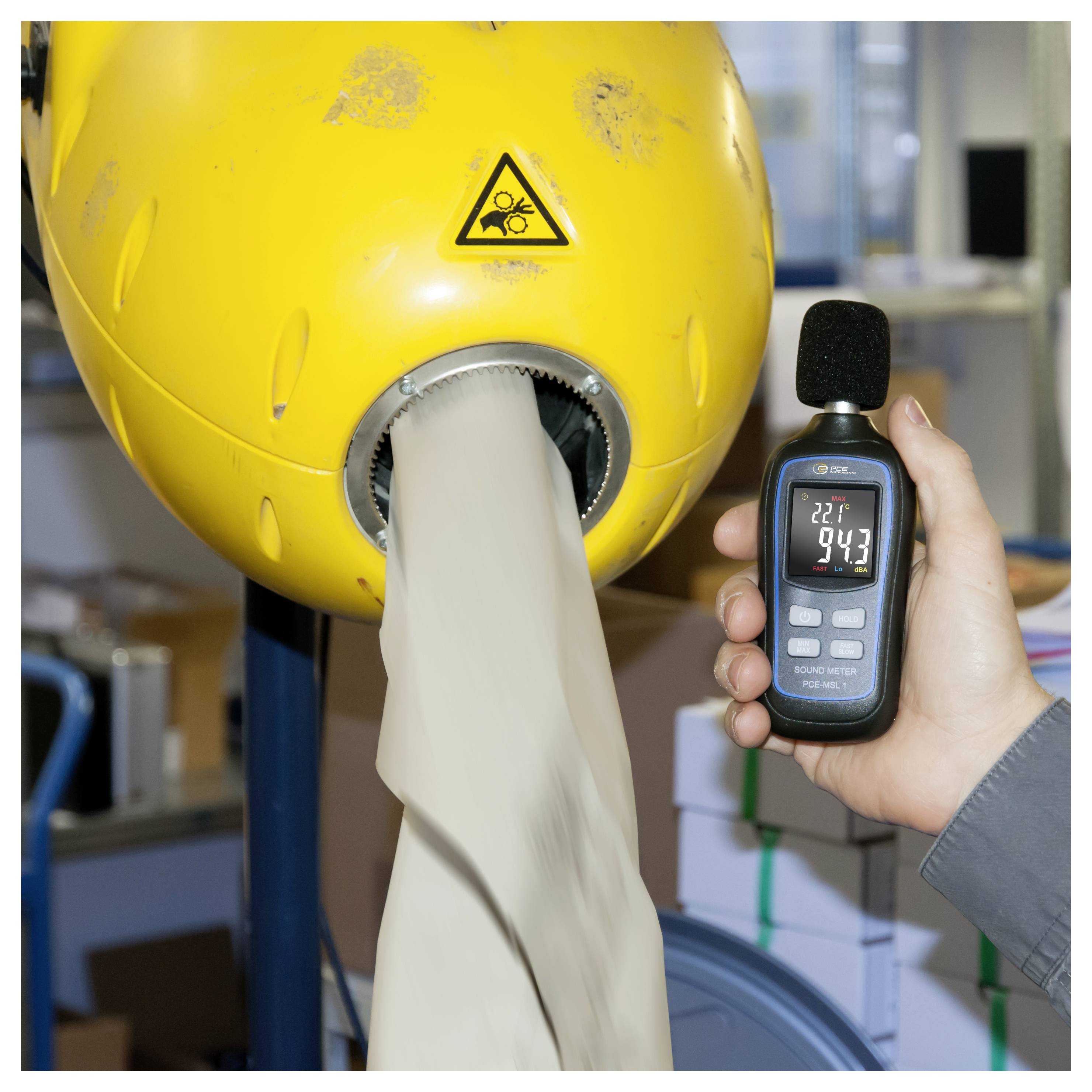 Handheld sound level meter measures noise from a yellow industrial machine with material feeding through an opening, showing a high reading.