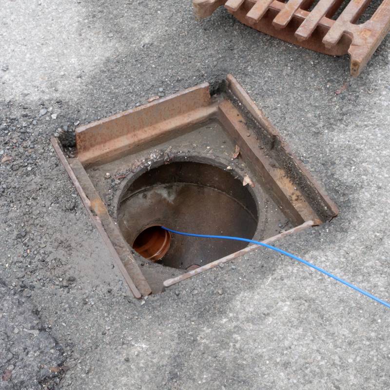 An open manhole cover with a visible sewage pipe and a blue cable inside, on an asphalt road.