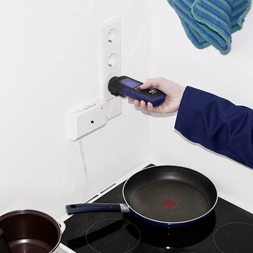 A person wearing a blue jacket is holding an energy consumption metre plugged into a socket. Alongside, a pot is sitting on a hob.