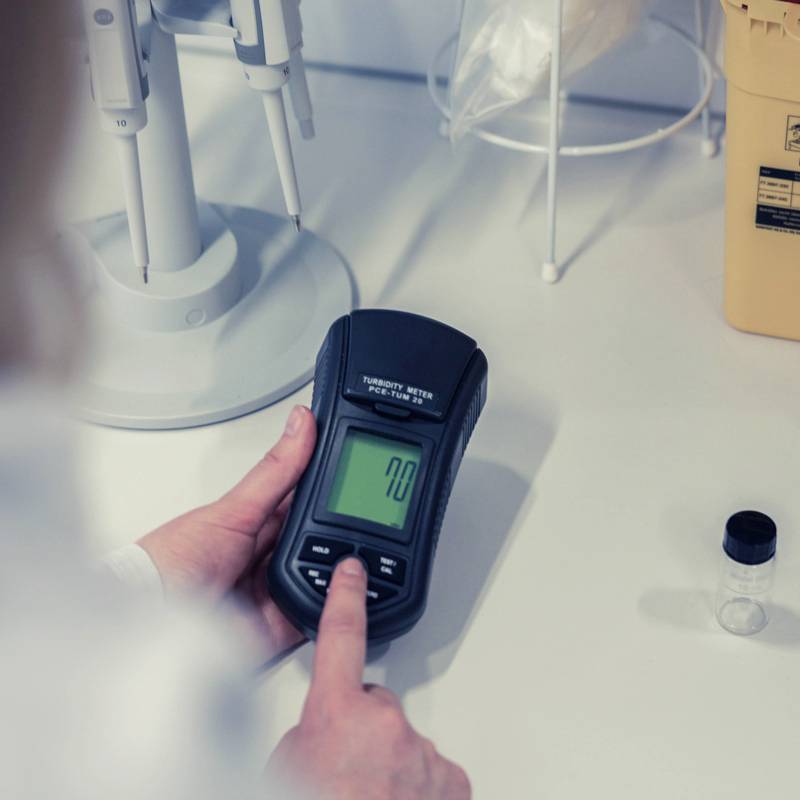 A hand is holding a measuring device with a digital display, showing the value '70'. Other laboratory equipment is placed on the table beside it.