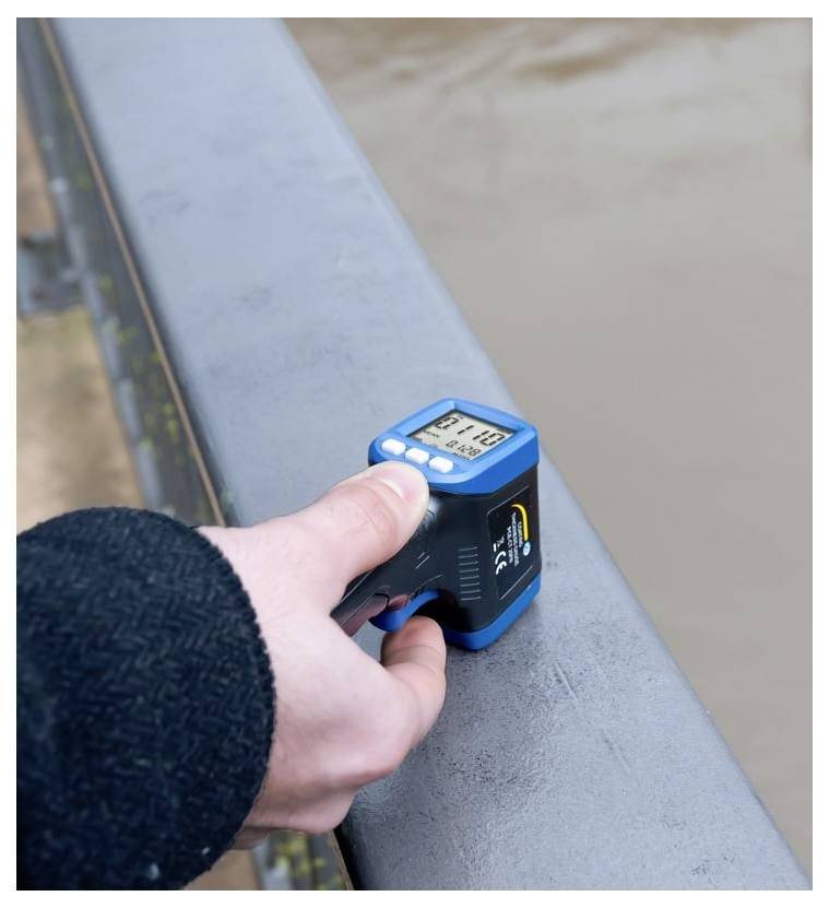 A hand holds a small digital device against a bridge railing, measuring conditions beside a river or canal.