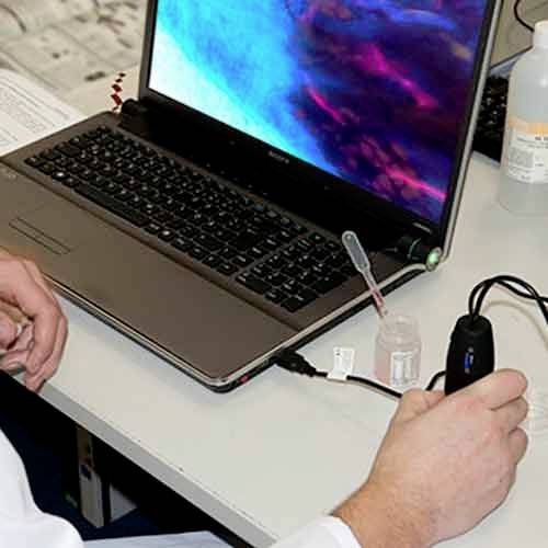 A person is conducting an experiment on a laptop, with a connected microscope and a small bottle of liquid.