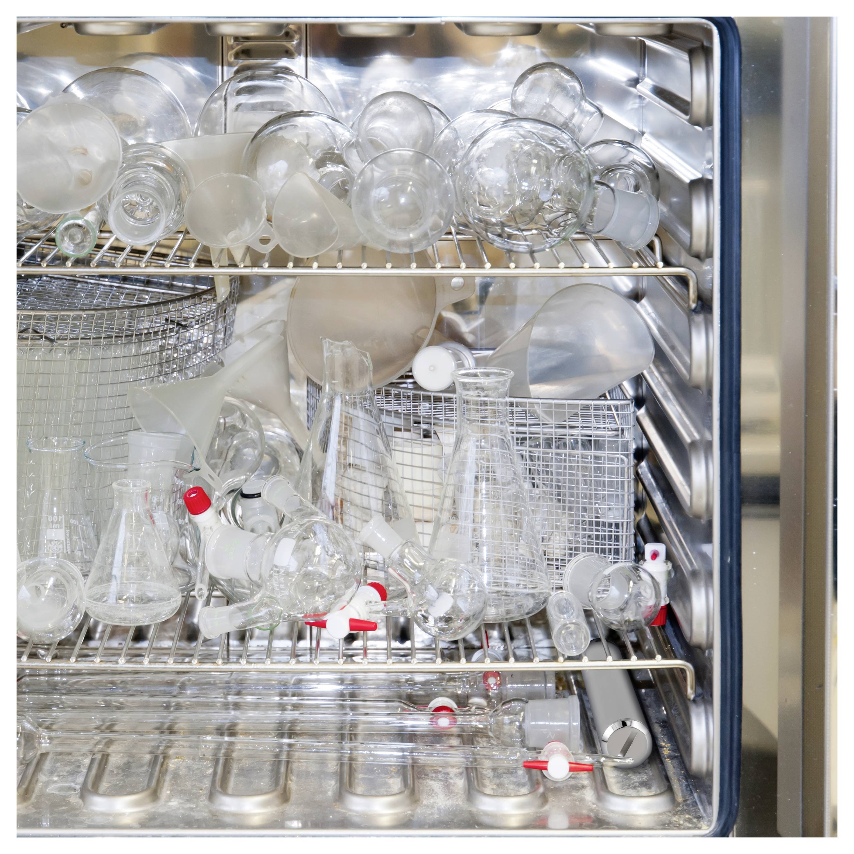 Dishwasher rack full of laboratory glassware and flasks drying inside an open stainless-steel chamber.