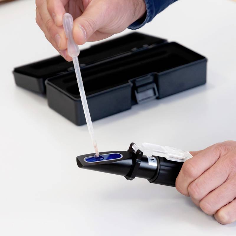 A person is using a hand refractometer to analyse a liquid. A black protective cover lies in the background.
