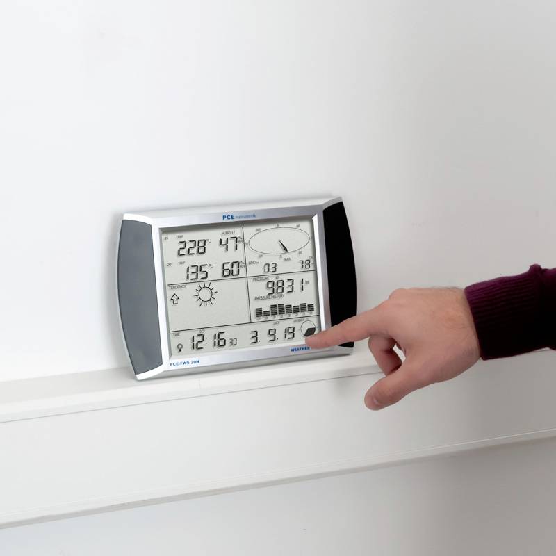 A person is pointing to an electronic measuring device with a digital display. Temperature, humidity, and pressure data are visible.