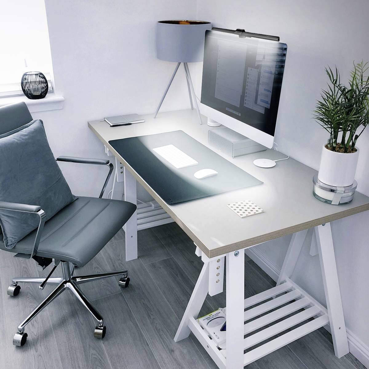 Modern workspace with a grey office chair, white desk, computer, lamp and plant, situated next to a window in the corner office.