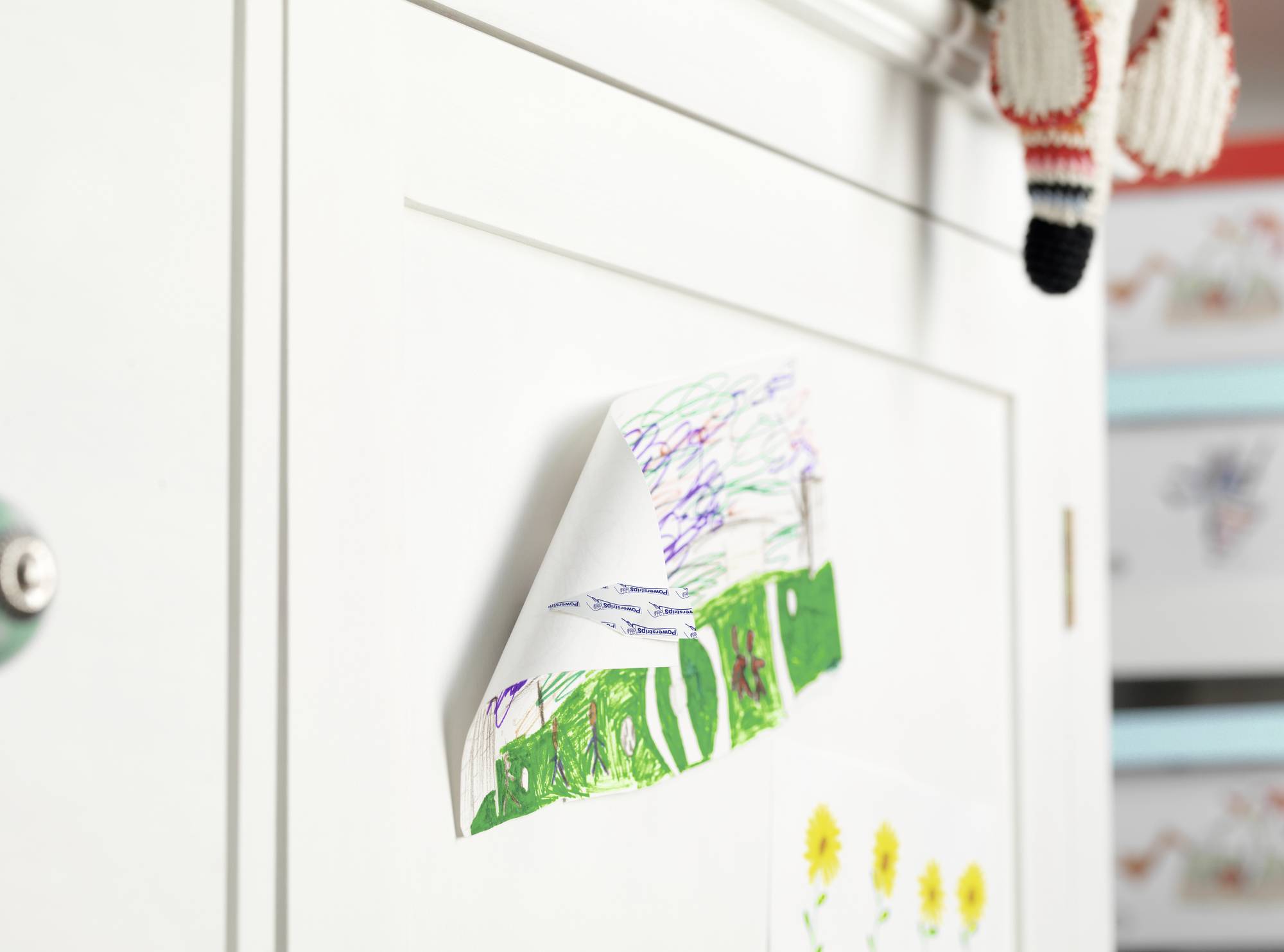 A colourful children's drawing of a house and a meadow is hung on a white cupboard. A knitted soft toy is hanging in the top right corner.