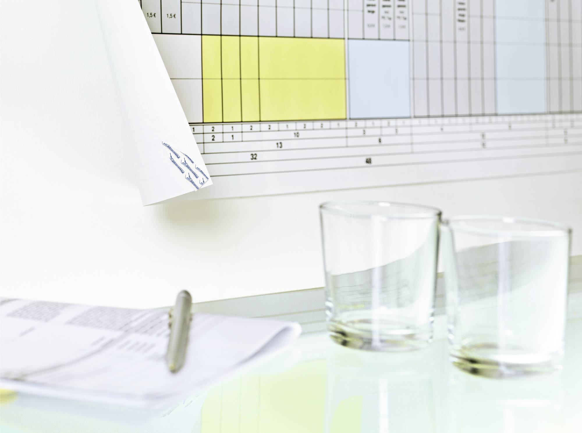 Two empty water glasses sit on a table next to an open notepad with a pen and a wall calendar in the background.