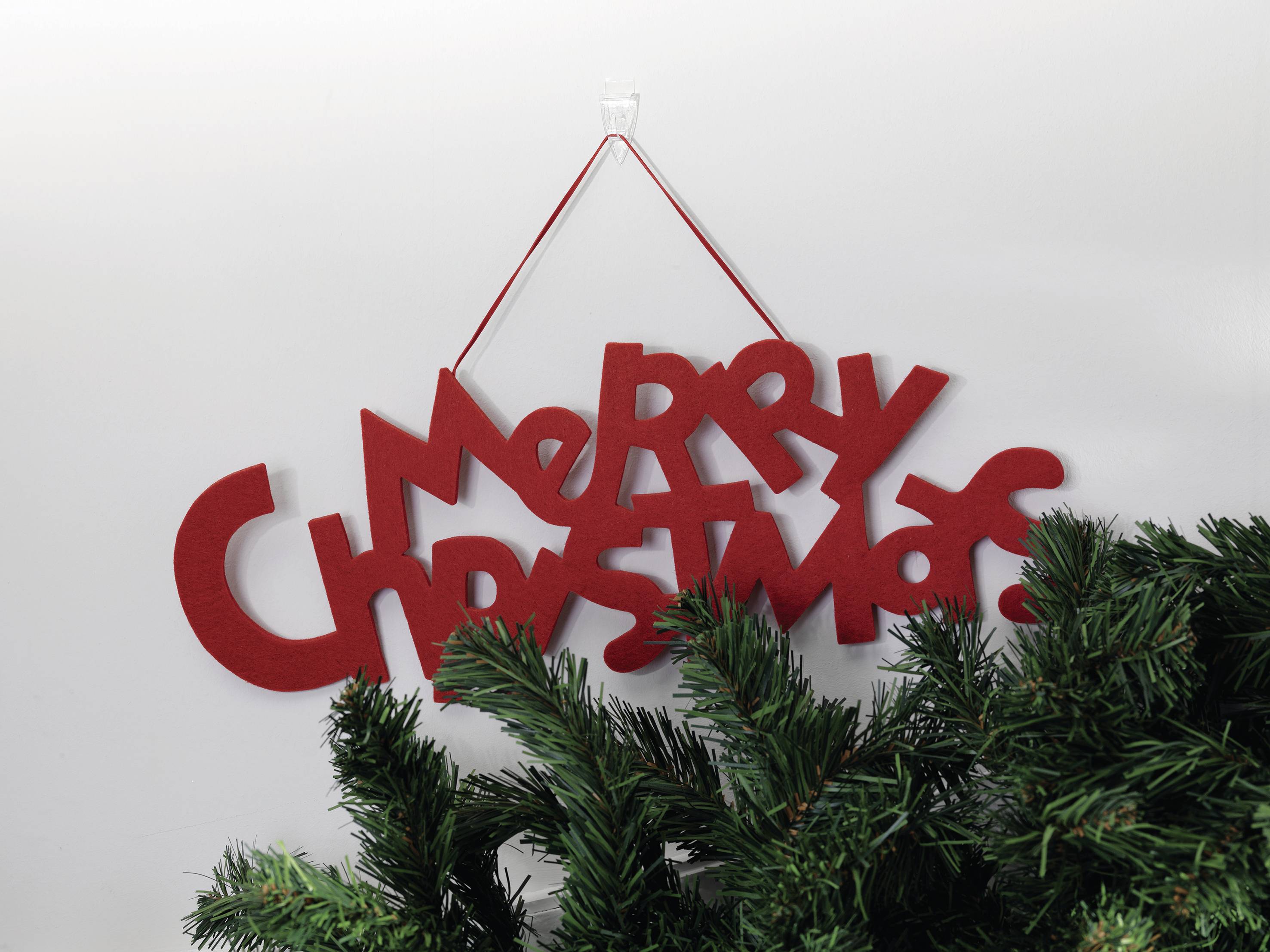 Red 'Merry Christmas' sign, hanging on a white wall. Pine branches placed underneath as decoration.