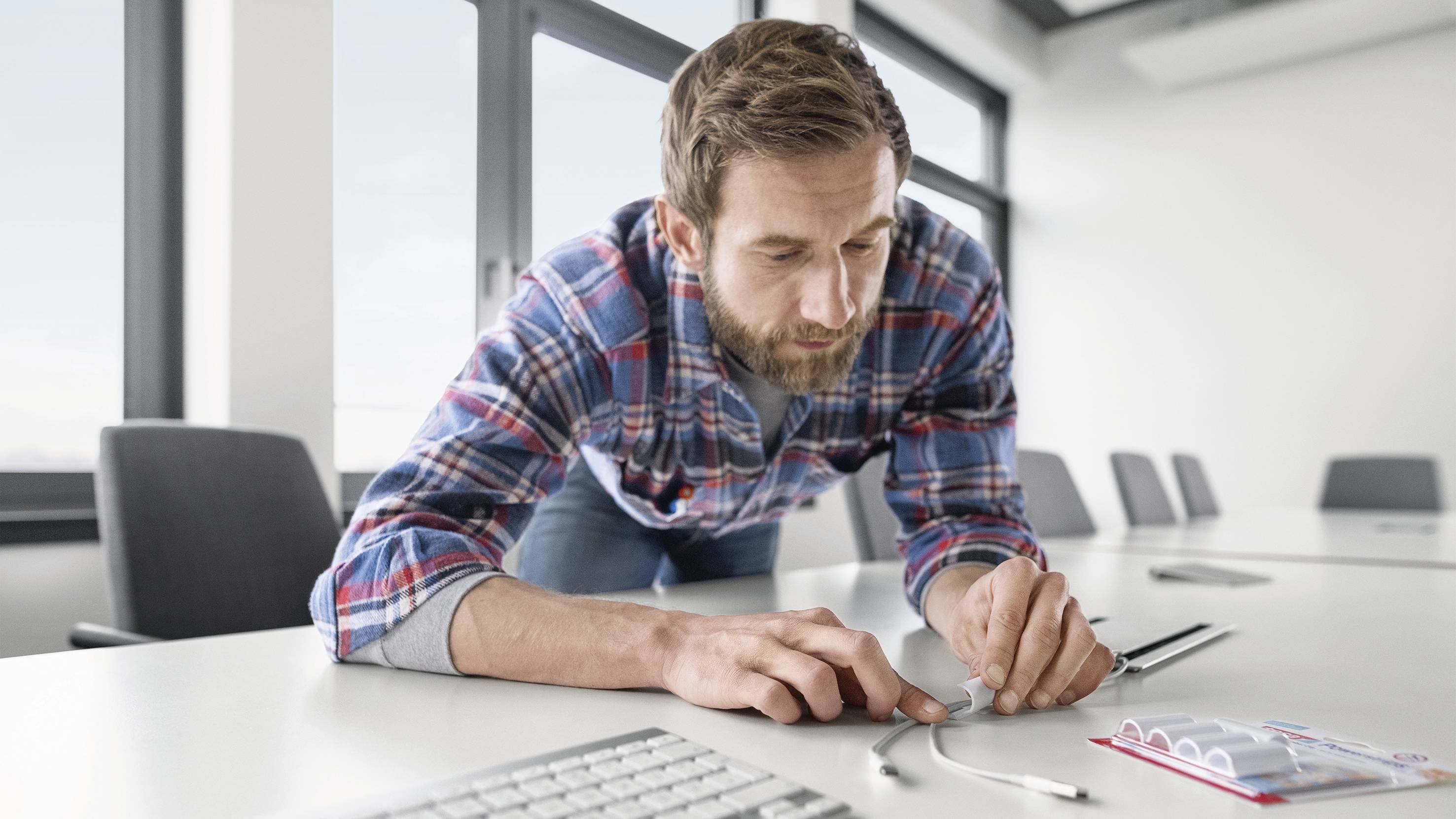 A man in a checked shirt is working attentively on cables at a table in an office. Windows and office furniture are visible in the background.
