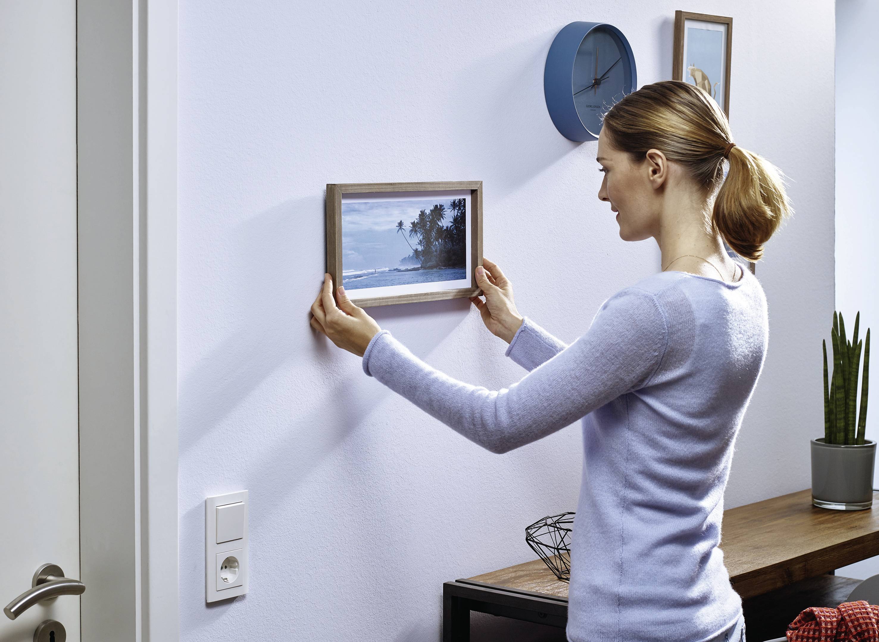 A woman is hanging a framed picture with a beach scene on the wall. In the background, a clock and plants can be seen.