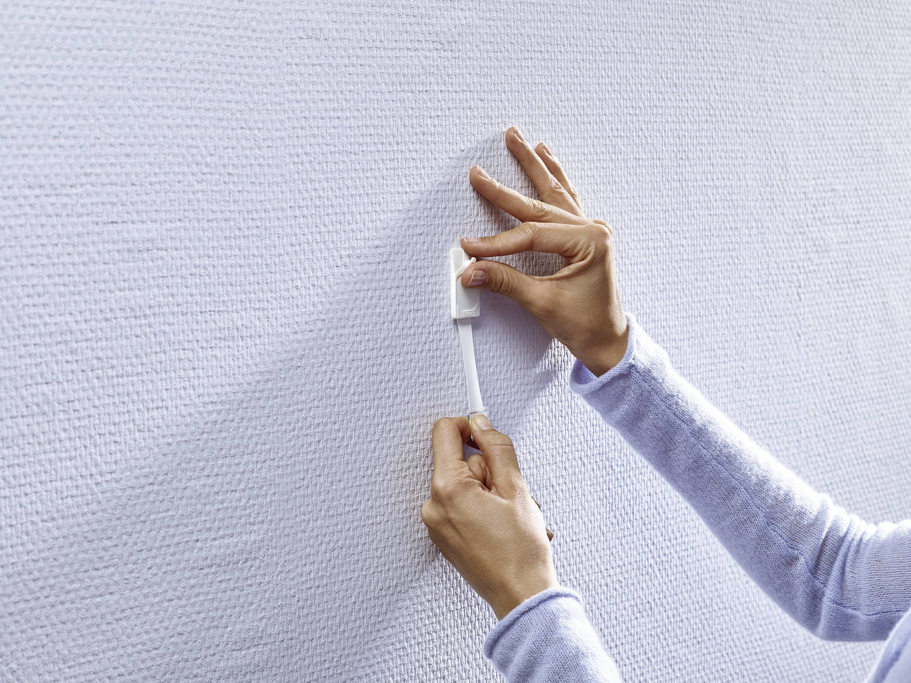 A person is attaching a self-adhesive hook-and-loop fastener to a white textured wall. Hands are holding the ends of the band.