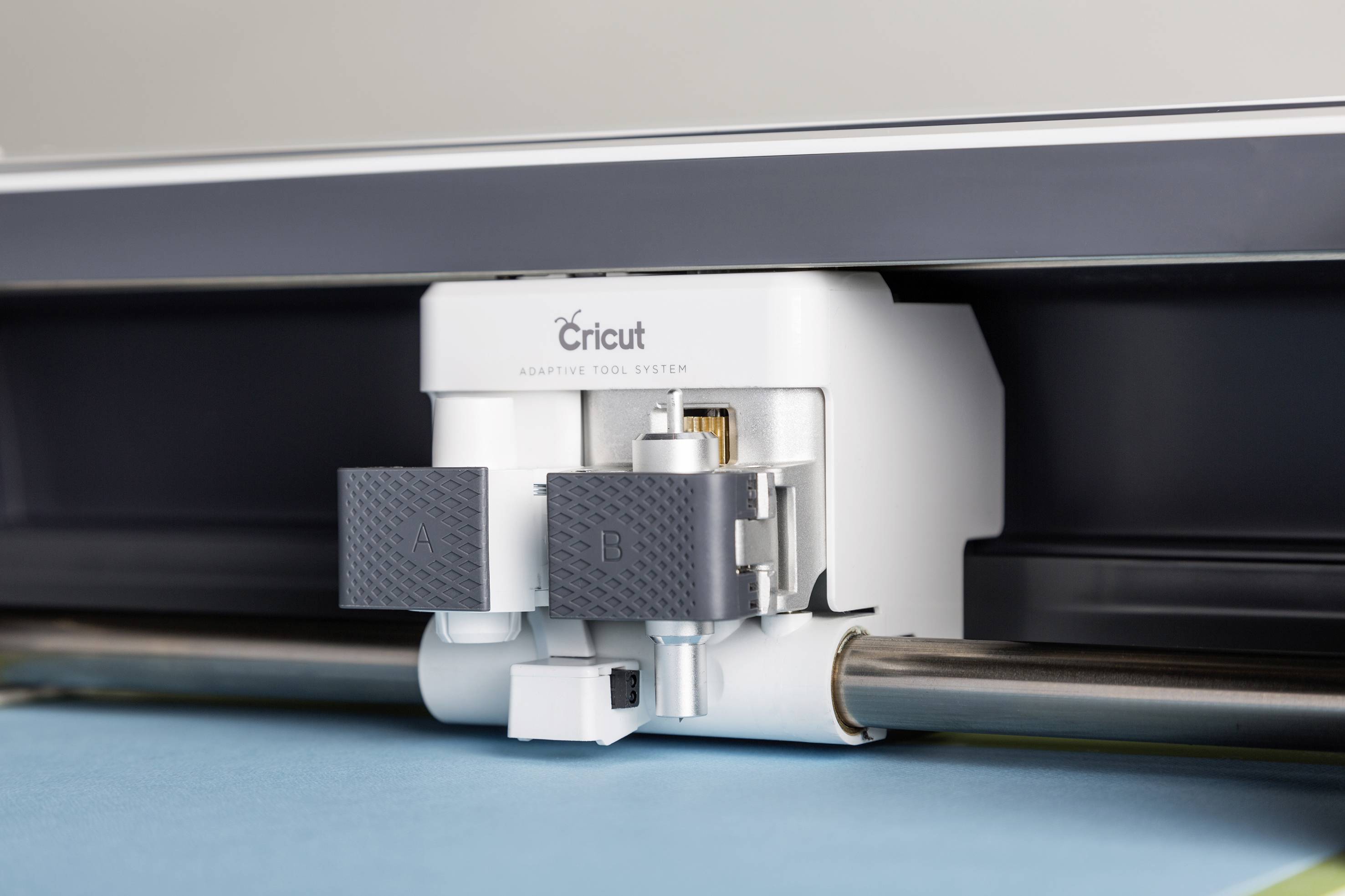 A cutting plotter cuts with precision on blue paper. The device has a white frame and two black cutting heads.