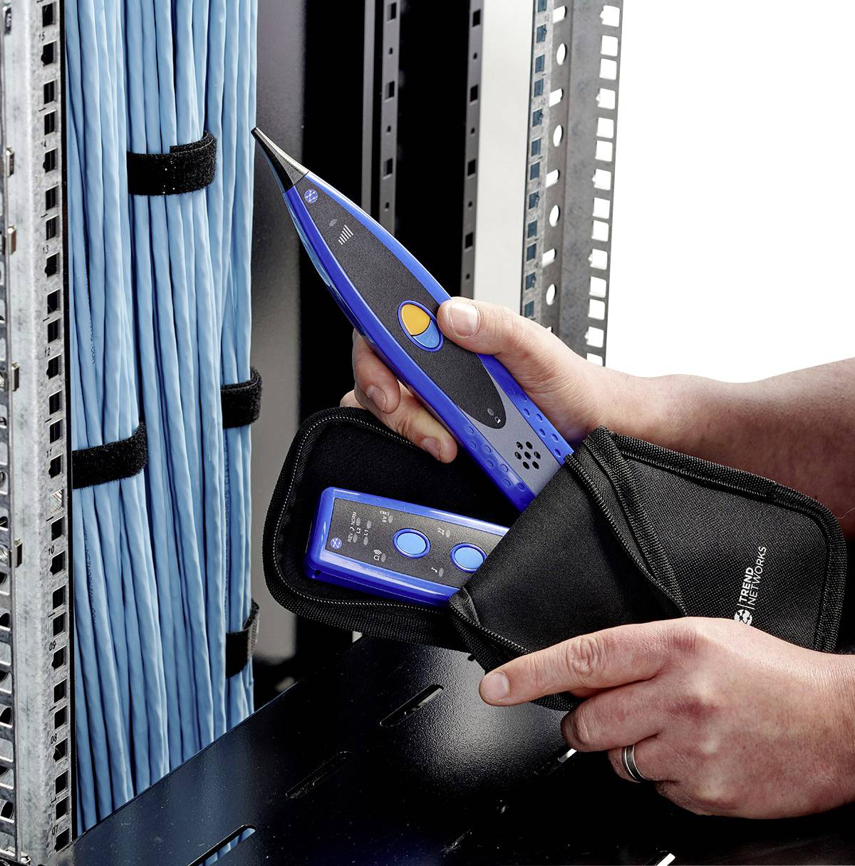 A person is holding a blue cable testing device next to an open server rack with blue cables, prepared for a network inspection.
