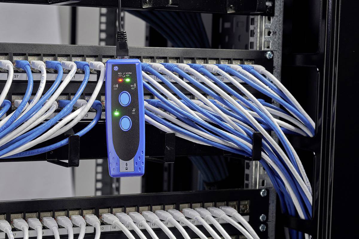 Server rack with connected blue and white Ethernet cables and a cable tester hanging on the cable harness with illuminated indicators.