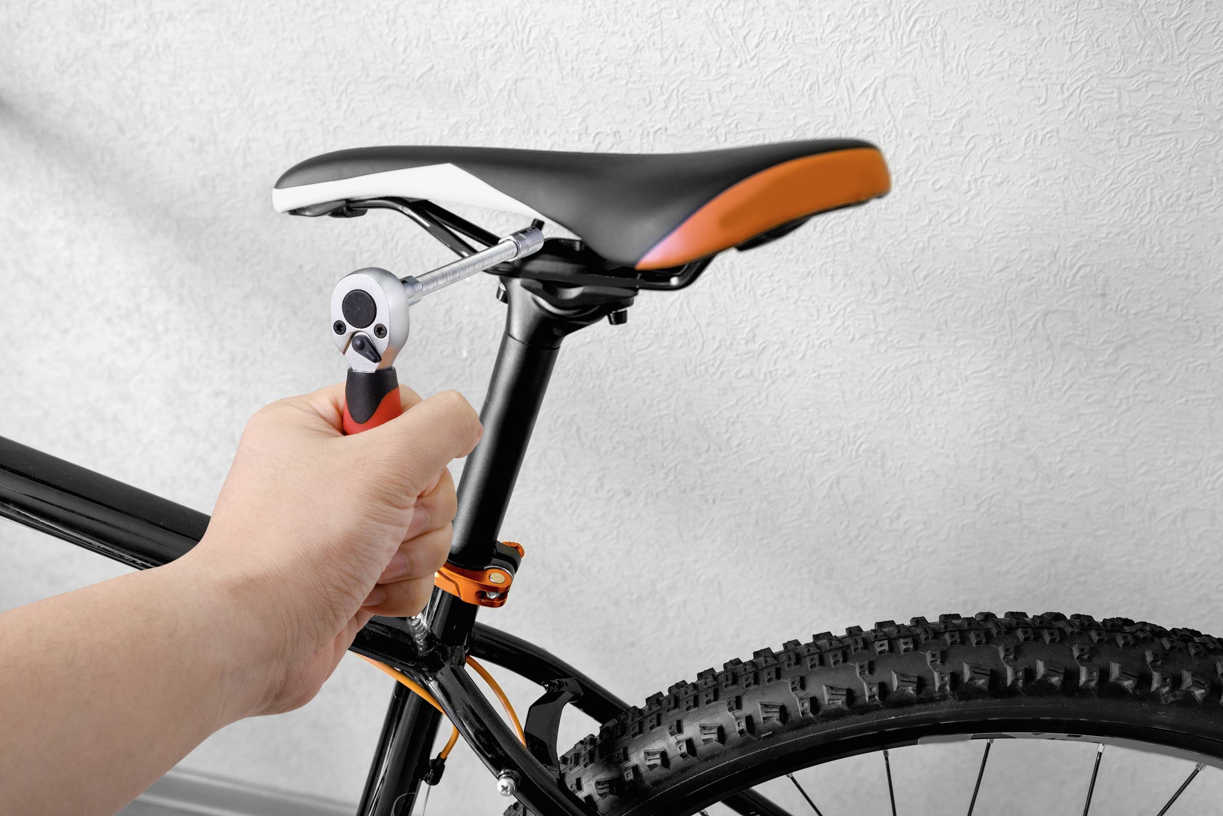A person is tightening the screws on a bicycle saddle with a spanner. The bicycle has a black frame and chunky tyres.