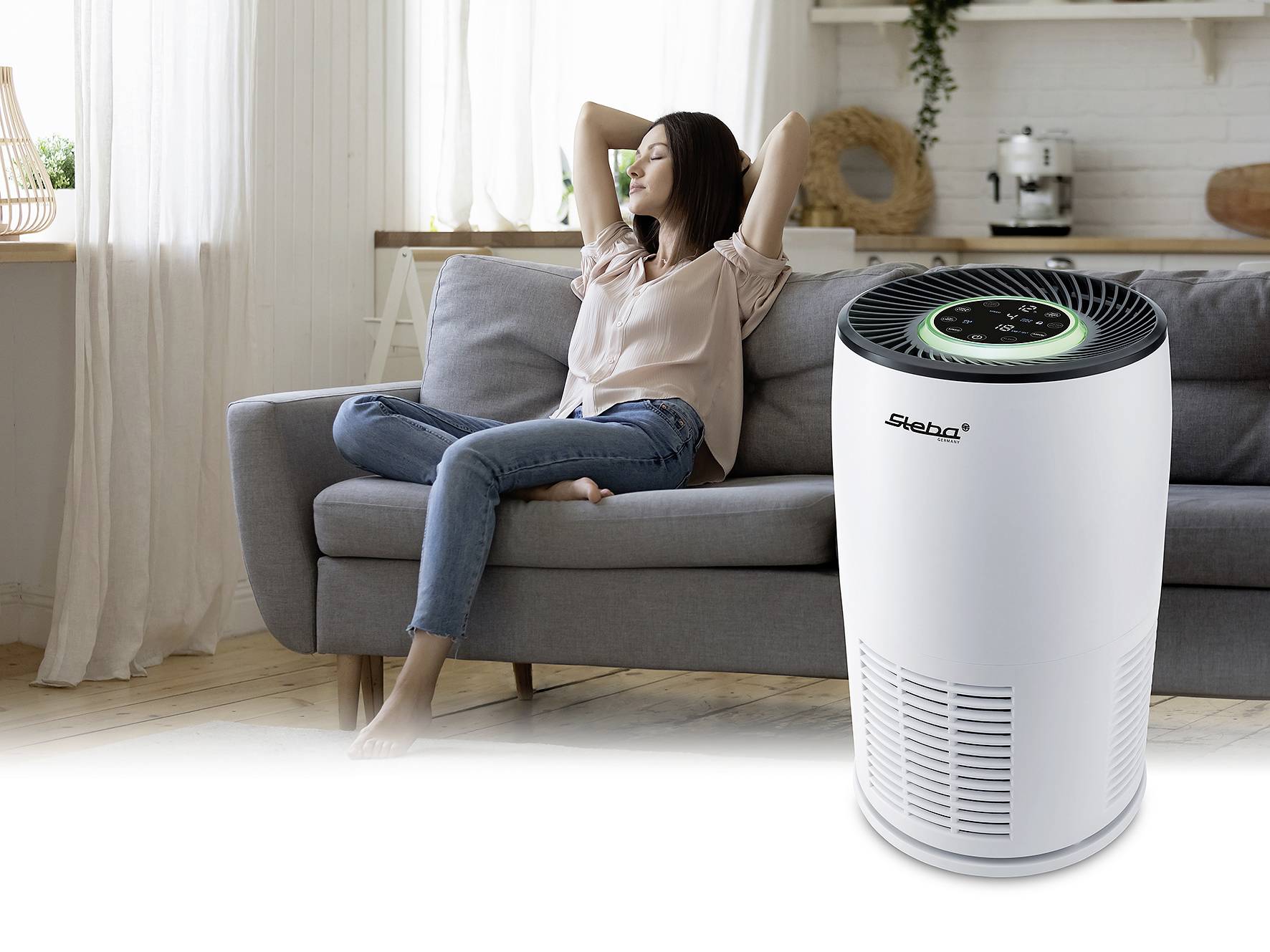 A woman relaxes on a sofa, with an air purifier featuring an illuminated control panel in the foreground. Living room furnishings are visible in the background.