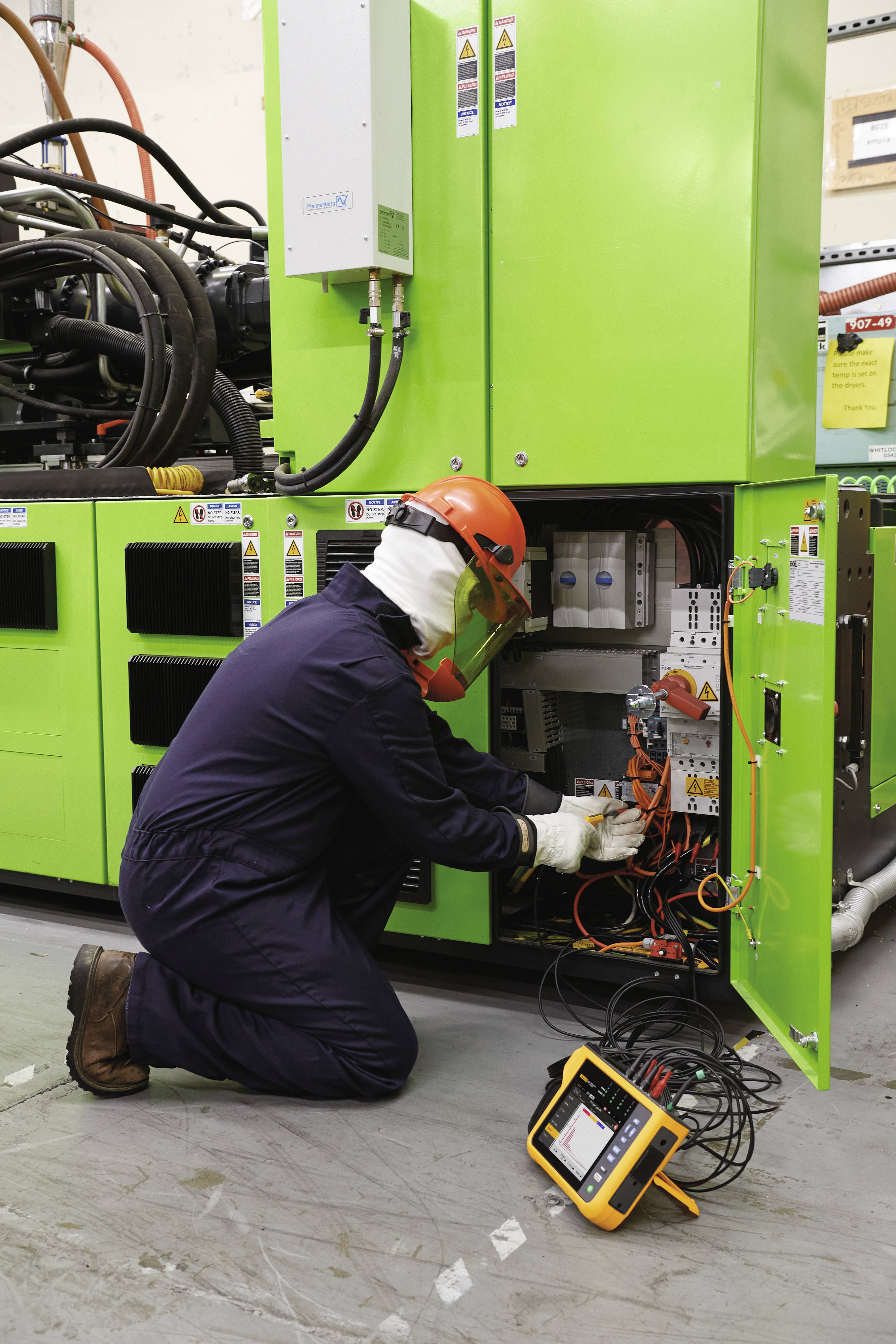 A technician in workwear and a hard hat is repairing a green machine. He is using a measuring instrument with connected cables.