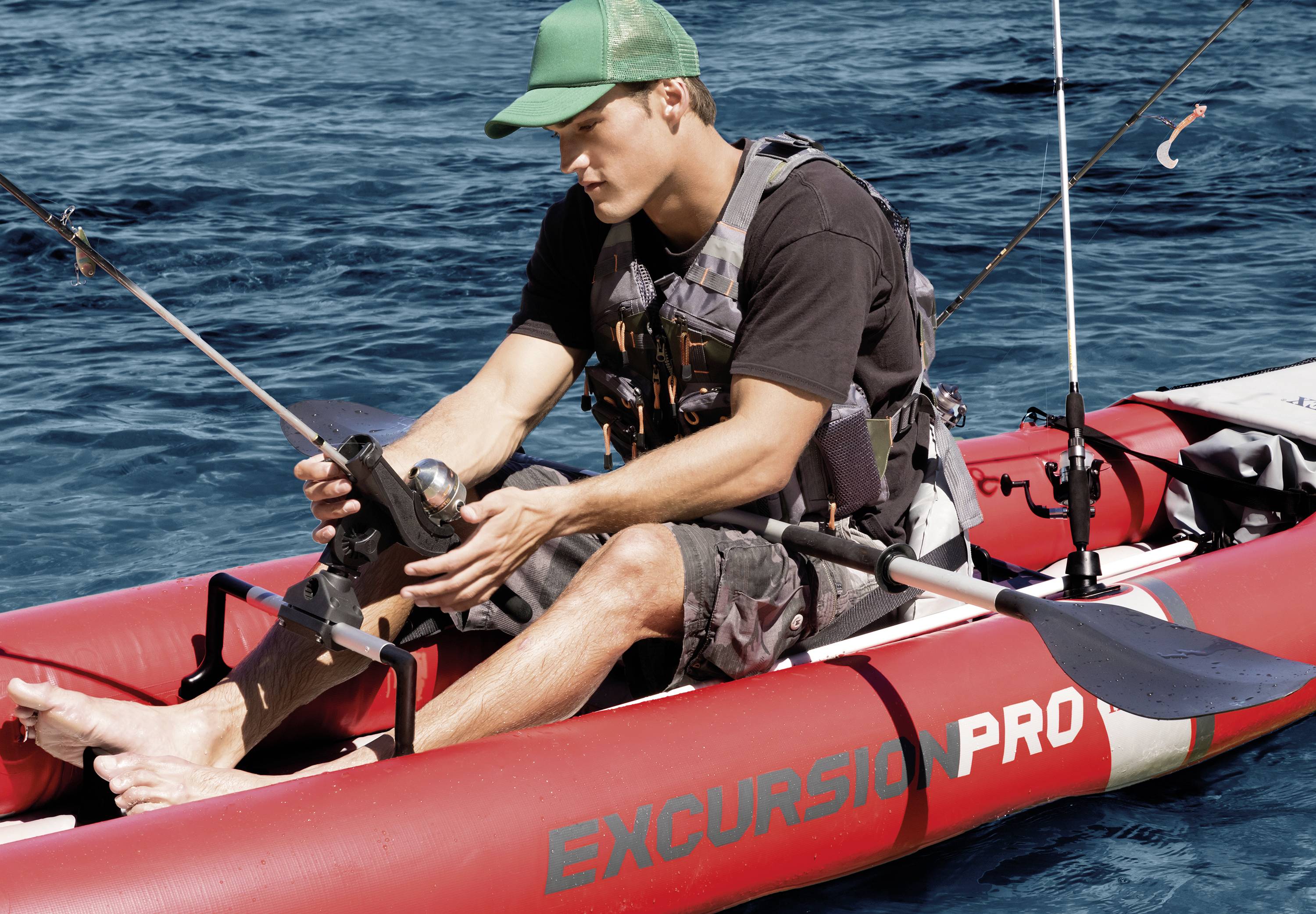 A man sits in a red inflatable dinghy on the sea, holding a fishing rod. He is wearing a green cap and a life jacket.