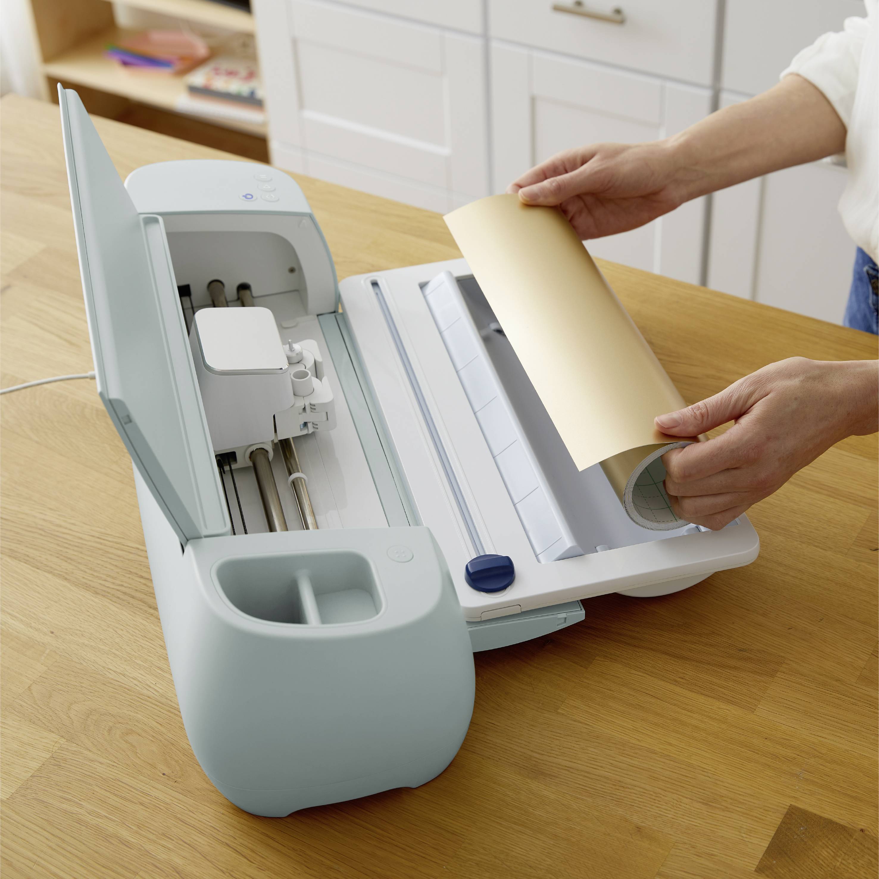 A person is placing a sheet of paper into a cutting machine situated on a wooden table in a modern, bright kitchen.