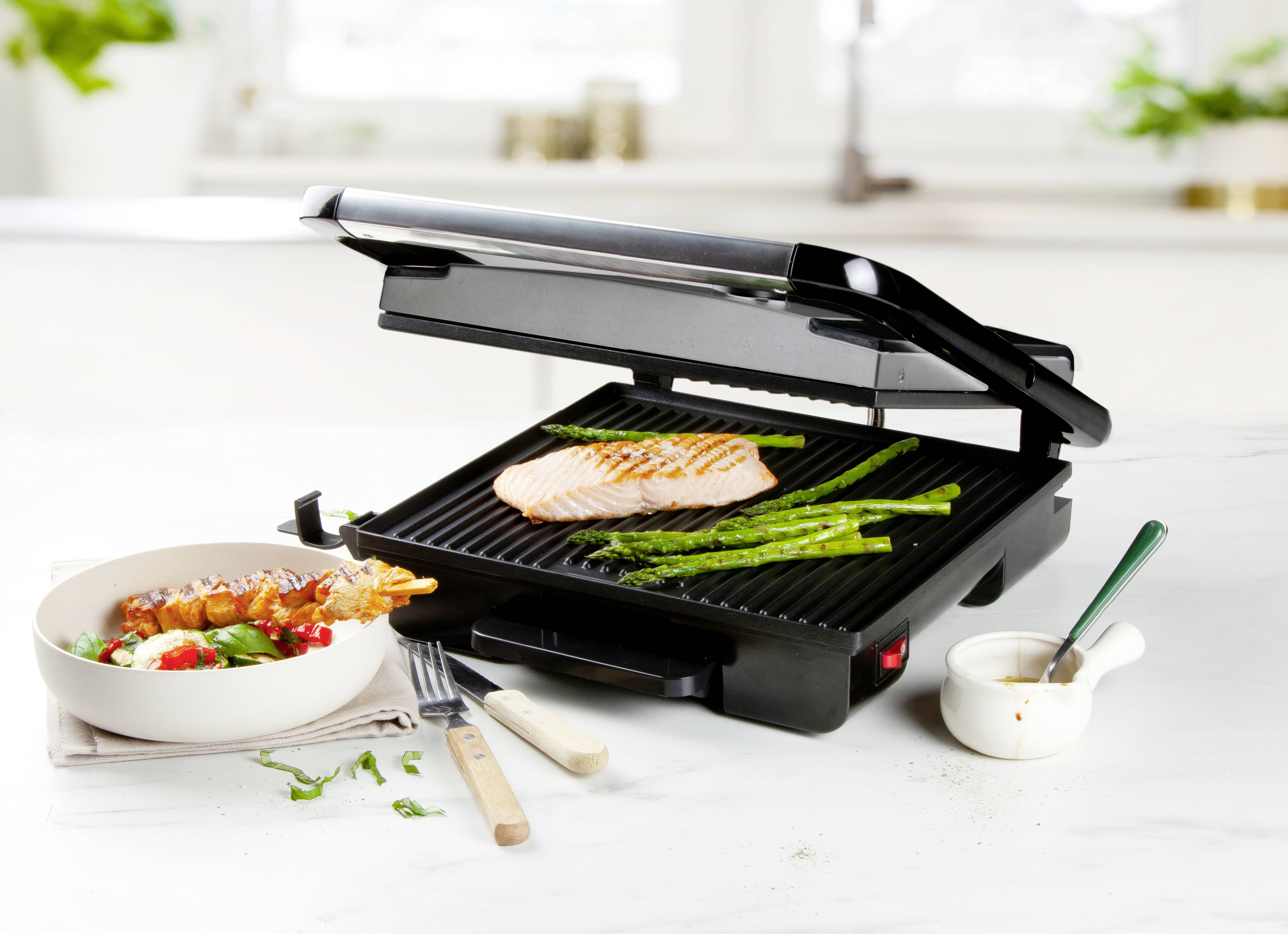 A contact grill on a kitchen worktop with its lid open. Salmon fillet and green asparagus are placed on it. A salad bowl is beside it.