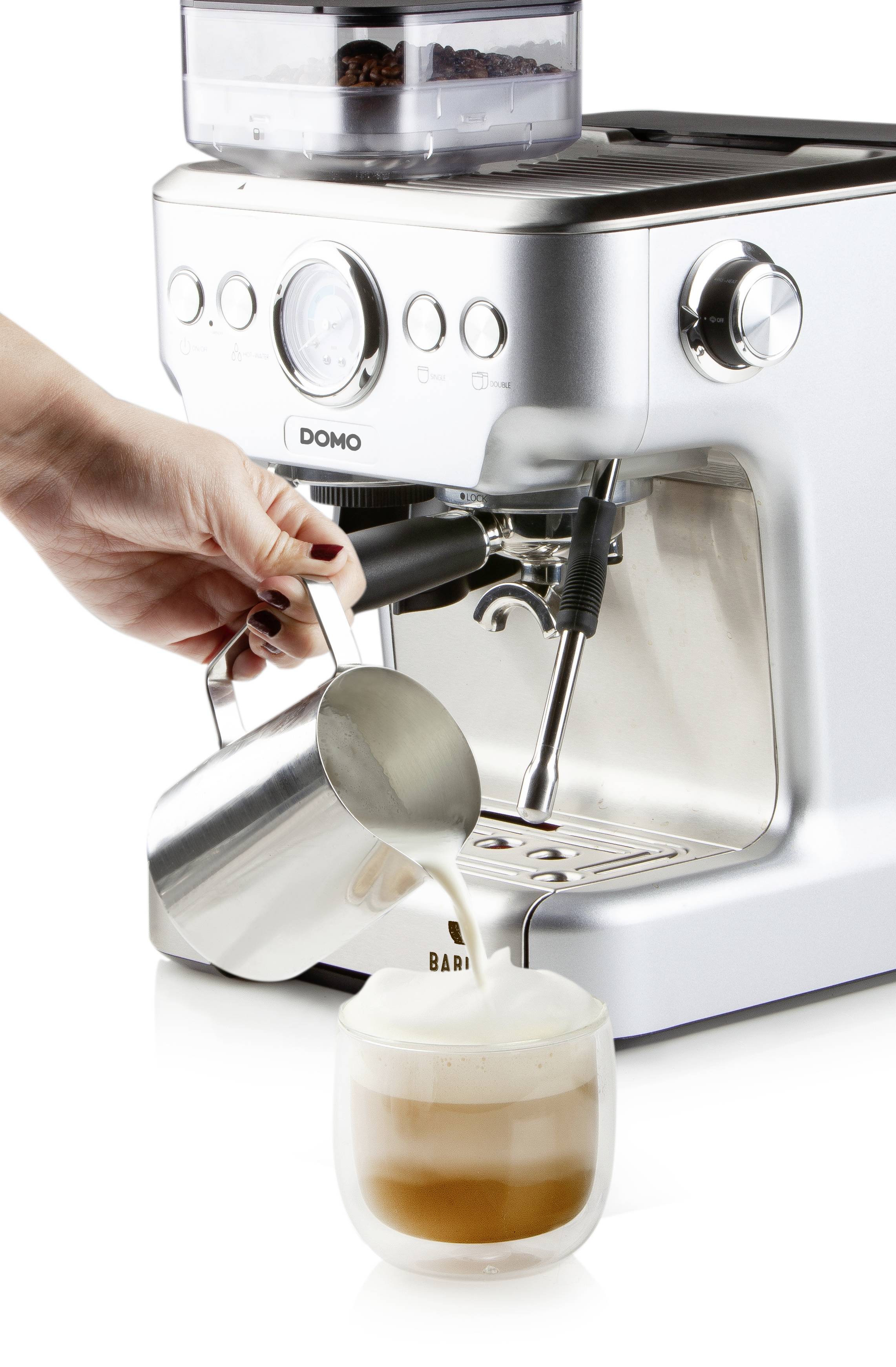 A person is pouring frothed milk into a cup of coffee next to a silver espresso machine.