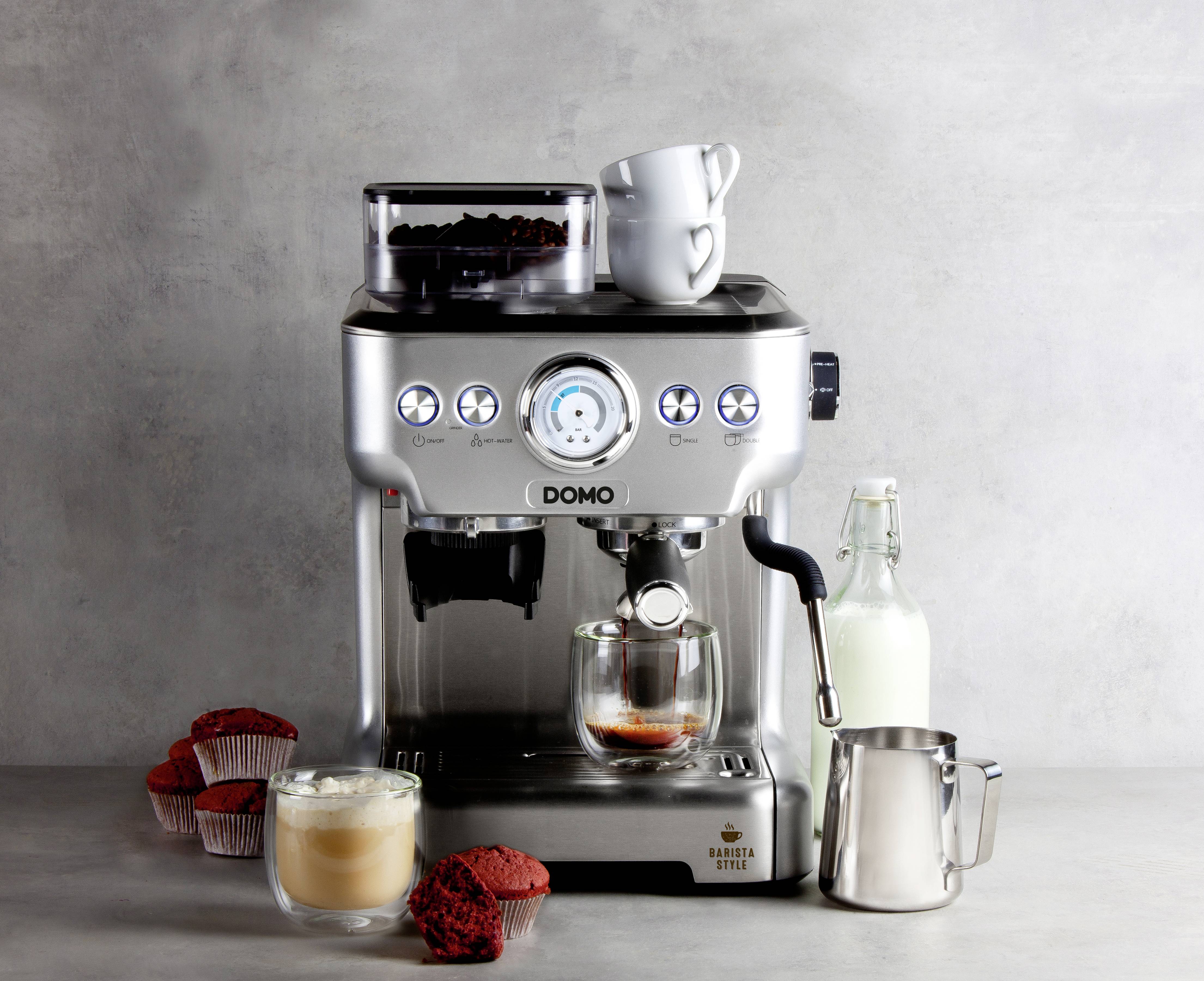 Espresso machine with steaming coffee in a glass, surrounded by cupcakes, milk bottle and milk jug on a grey background.