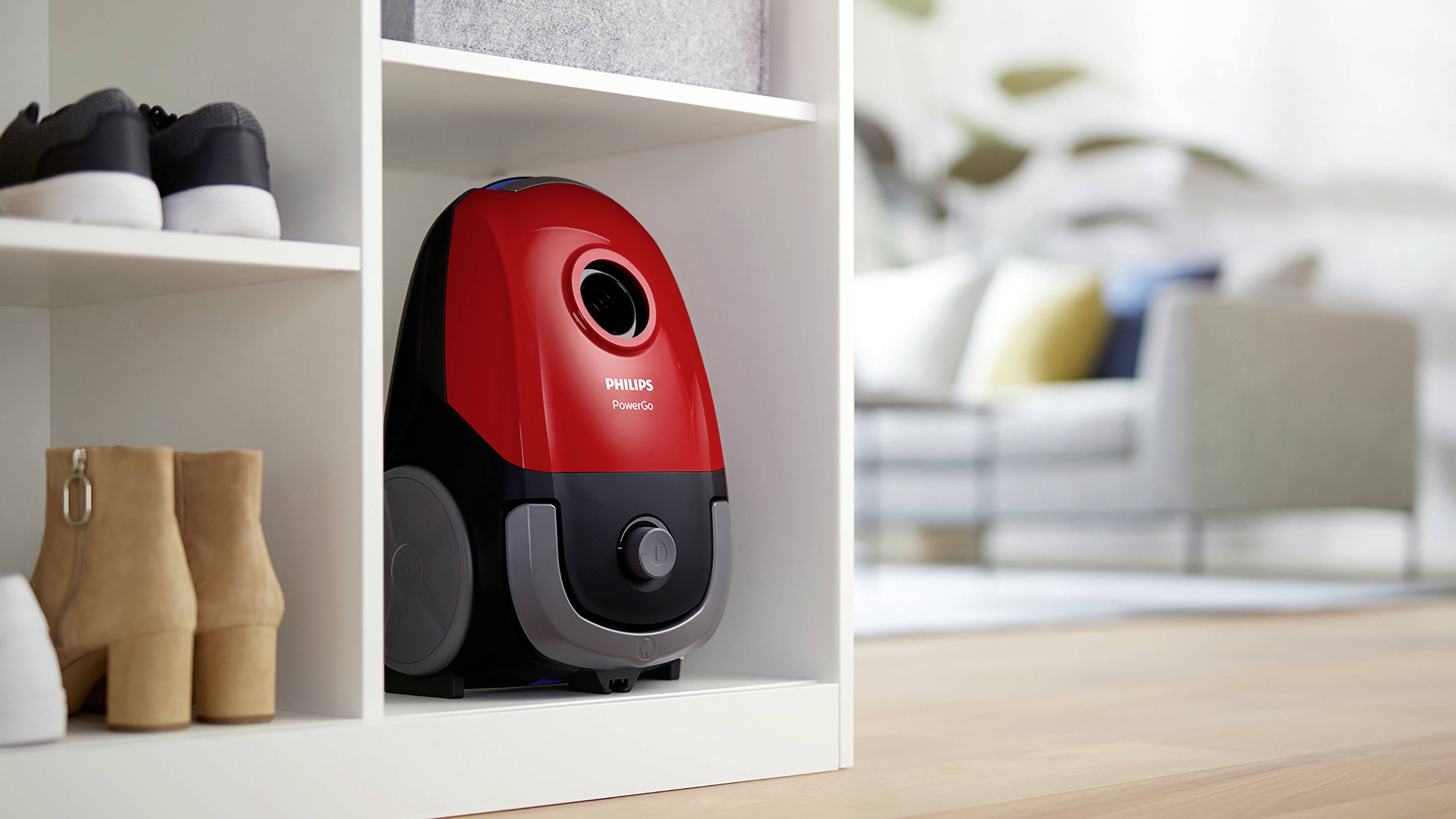Vacuum cleaner on a shelf, next to shoes, in the living room. The vacuum cleaner is red and black. The room displays a modern interior design.