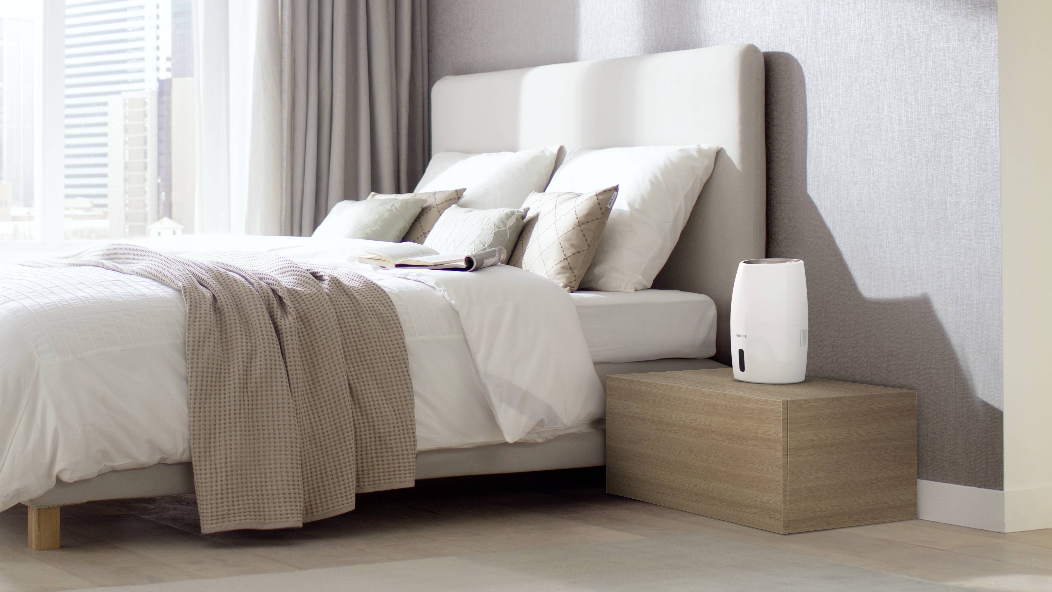 Bedroom with a double bed, numerous pillows, duvet and bedside table with a white air humidifier, next to tall windows with curtains.