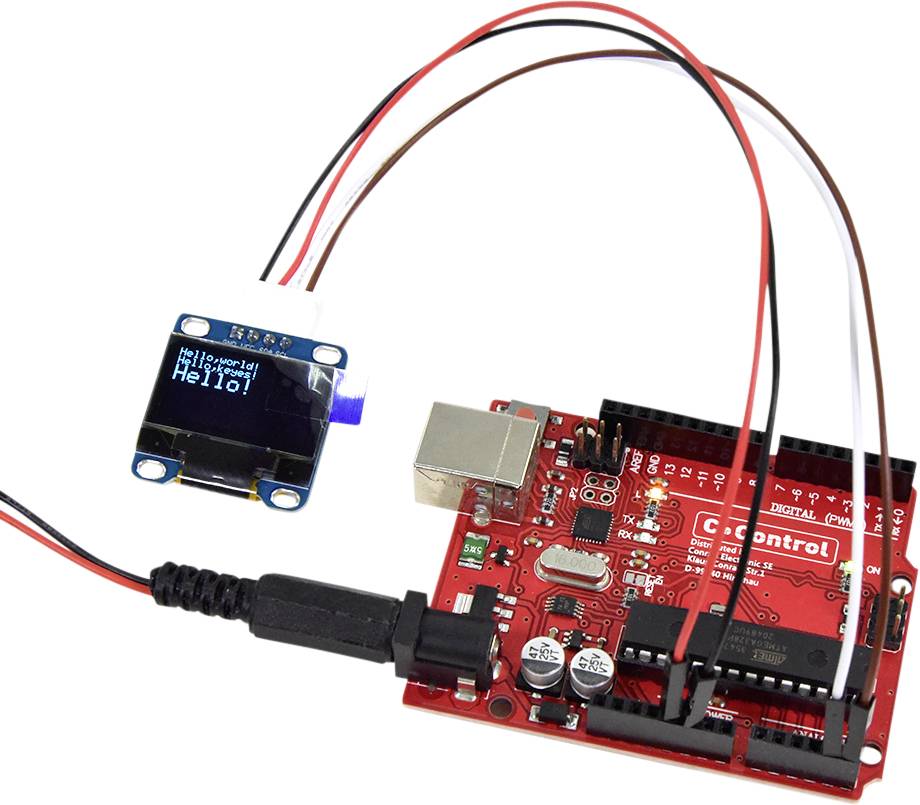 A red electronic circuit board is connected to a small display showing 'Hello!'. Several cables are connected.