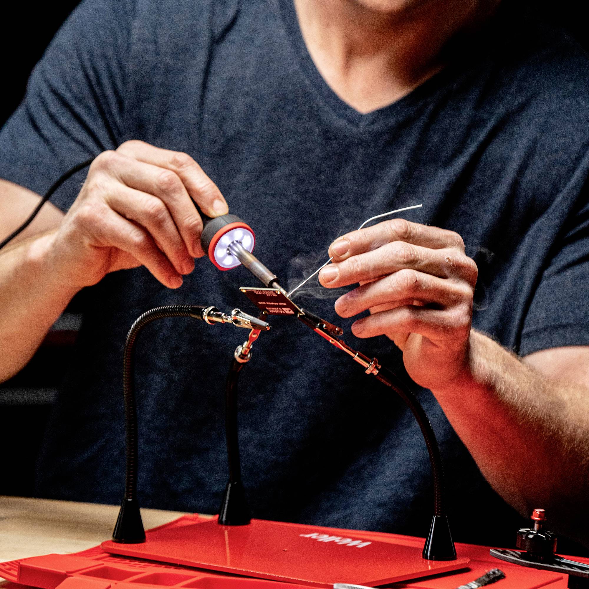 A person is soldering a small electronic component on a red table. They are holding a soldering iron and solder.